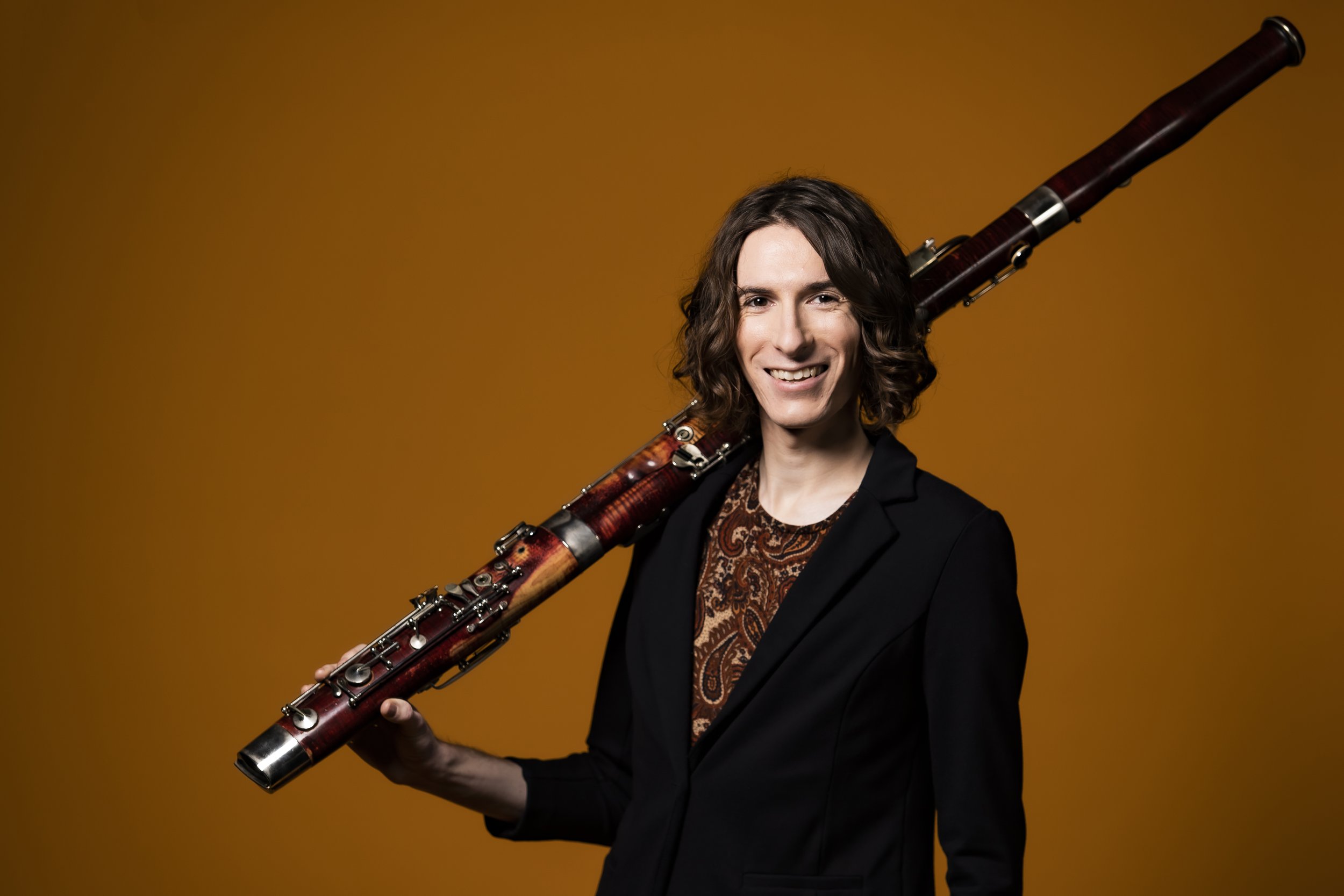 Andy Sledge holding their bassoon casually on their shoulder, smiling into the camera in front of an orange background.