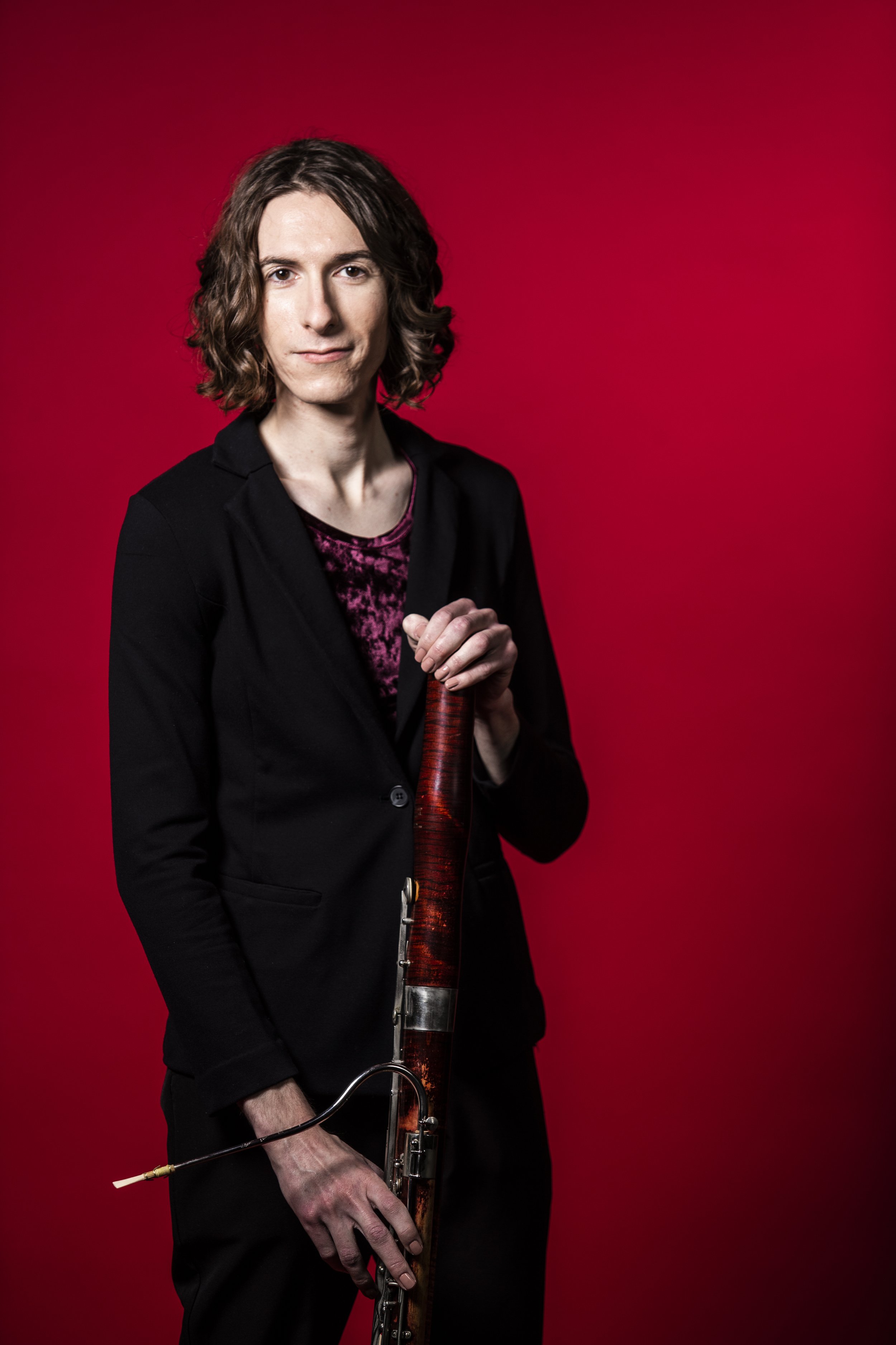 Andy Sledge holding a bassoon upright, acknowledging the camera directly in front of a red background.