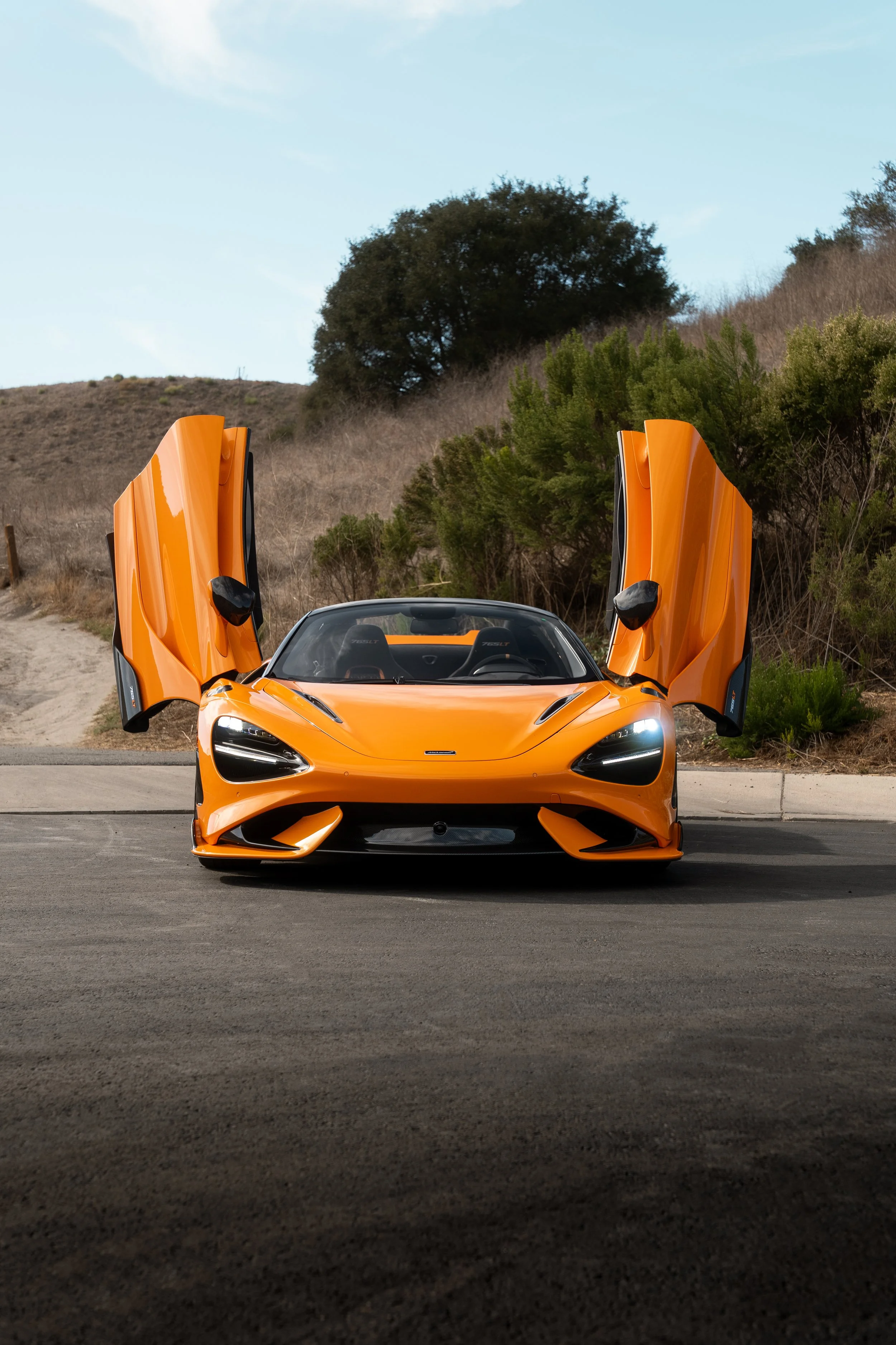 Orange McLaren supercar with butterfly doors raised, parked outdoors on a paved road.