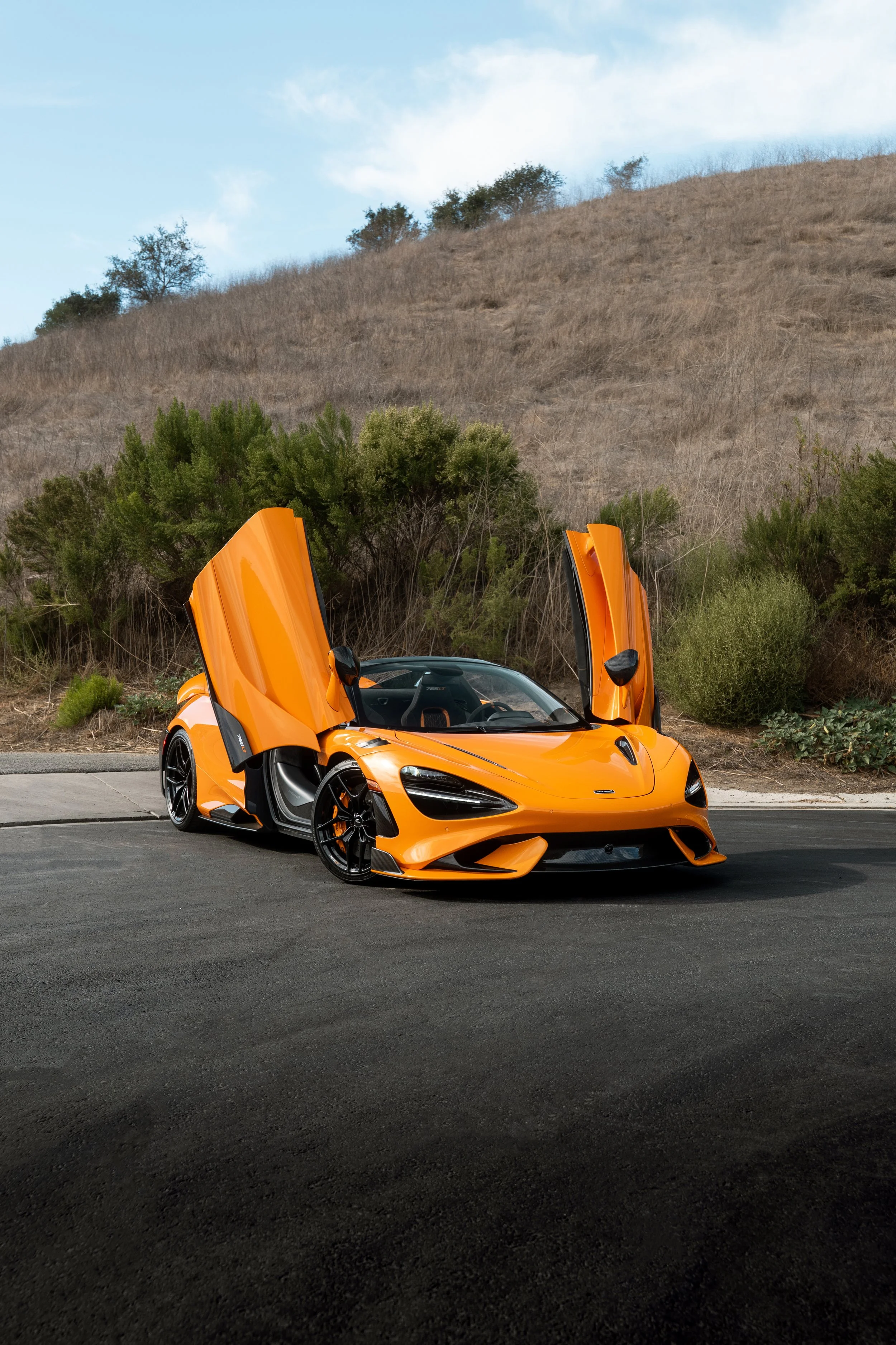 Orange McLaren sports car with butterfly doors open parked on a road beside a dry grassy hillside
