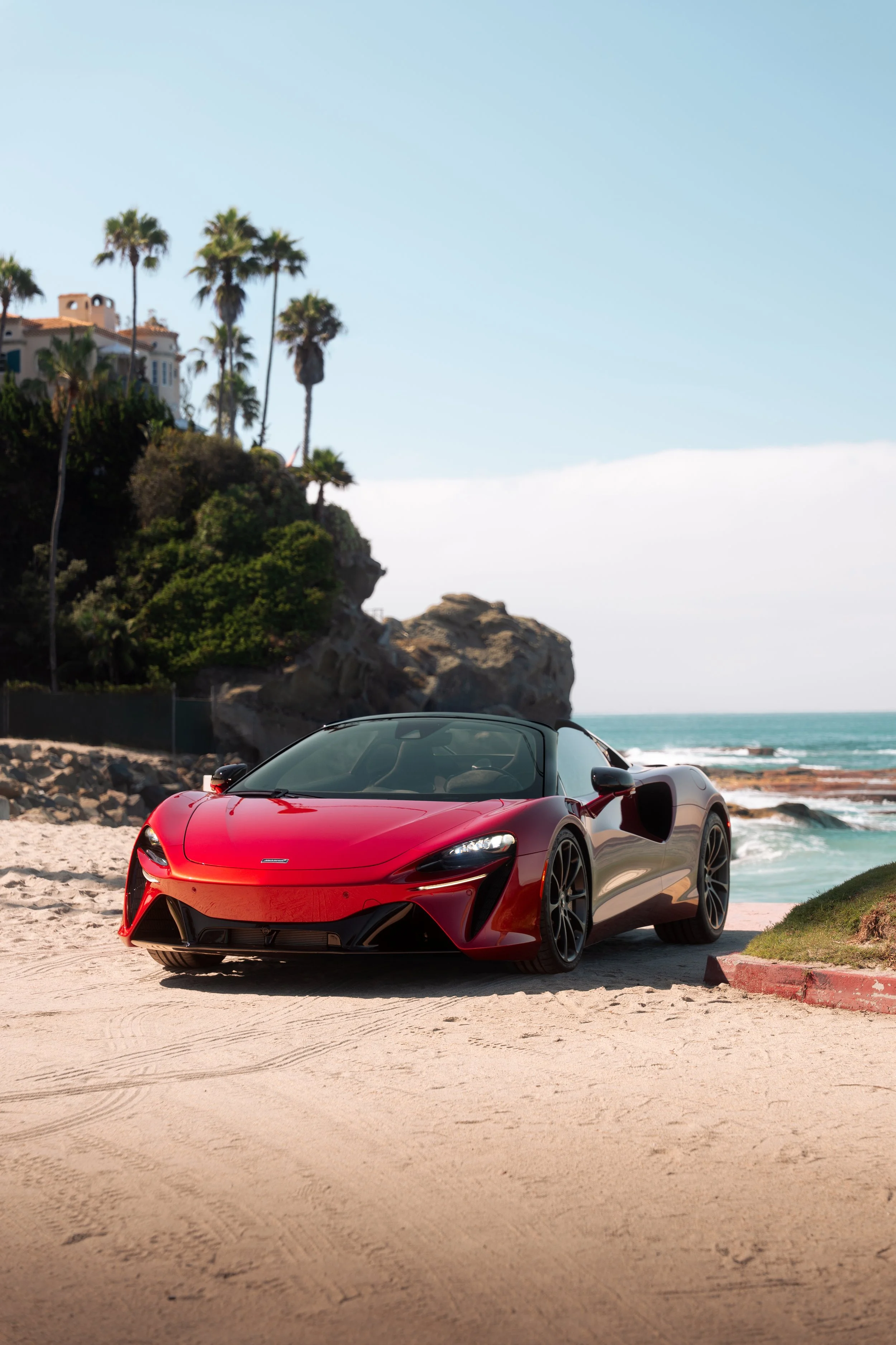 Red sports car parked on a sandy beach beside ocean waves, palm trees, rocks, and a coastal villa.