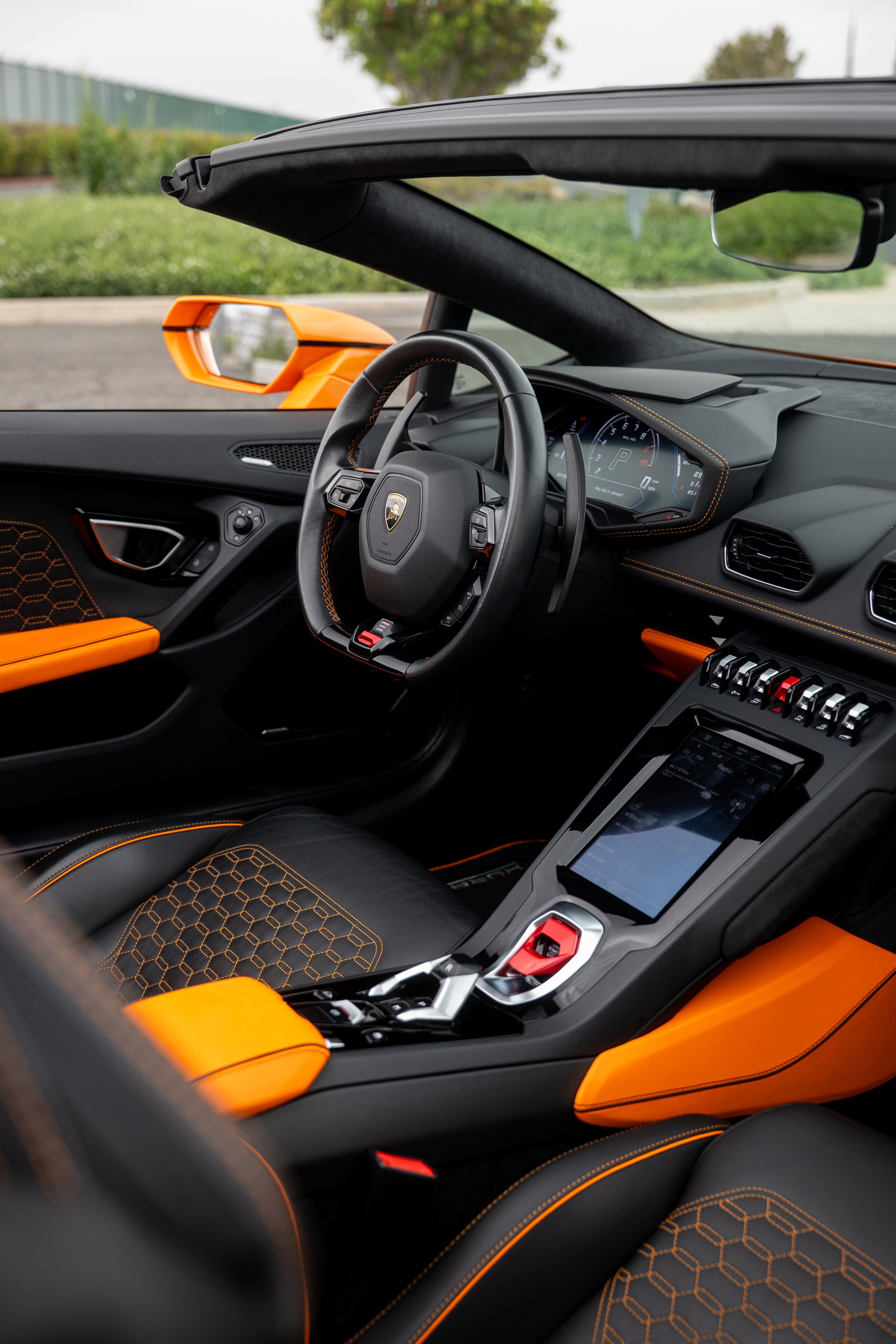 Interior of an orange Lamborghini with black leather seats, sport steering wheel, digital dashboard, and center console.