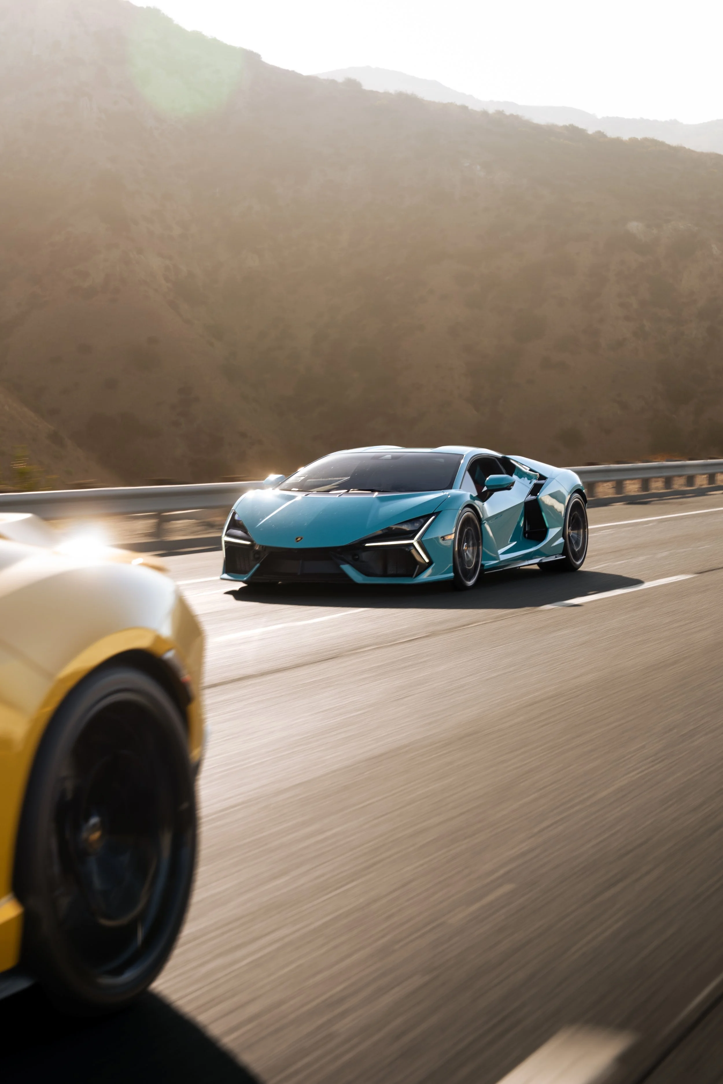 Turquoise Lamborghini supercar speeding on a mountain highway beside a blurred yellow sports car