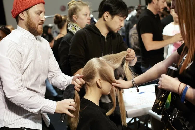 A woman hairstylist is blow-drying and styling a woman's hair at a busy hair or beauty event, with multiple people in the background.