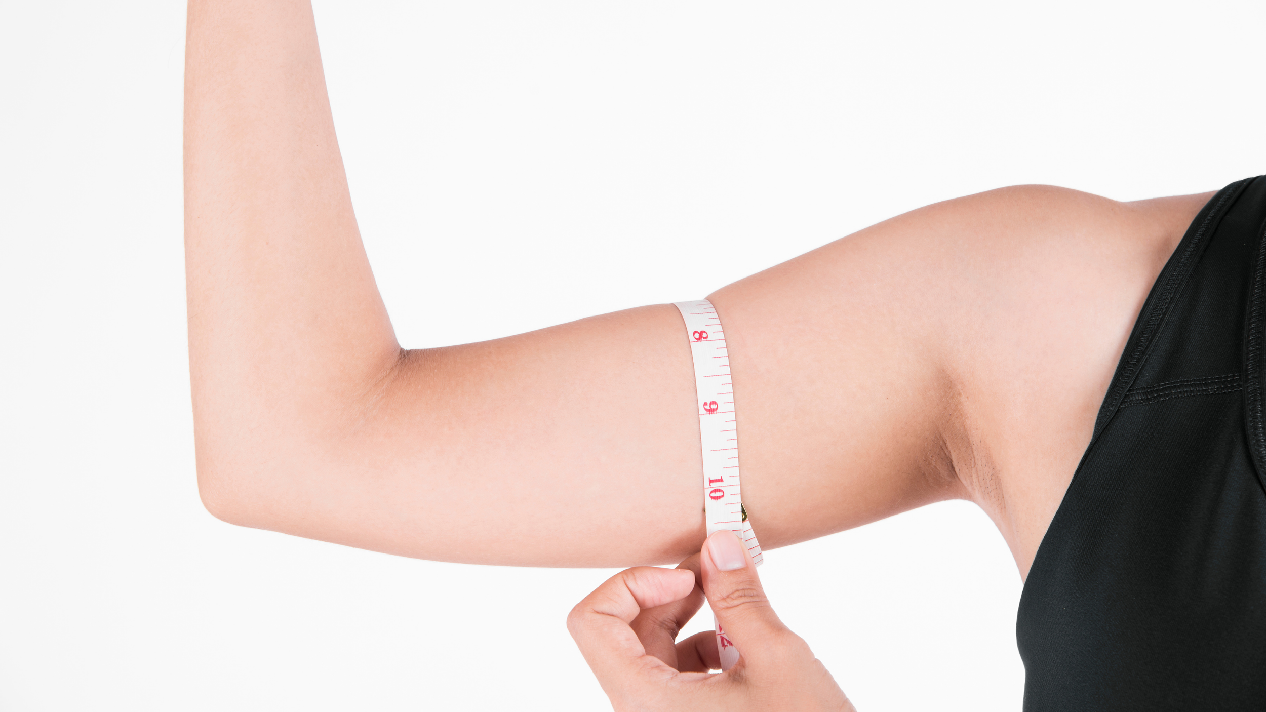 A person measures their upper arm with a tape measure on a white background. The arm is bent, and they wear a black sleeveless top, conveying focus and fitness.