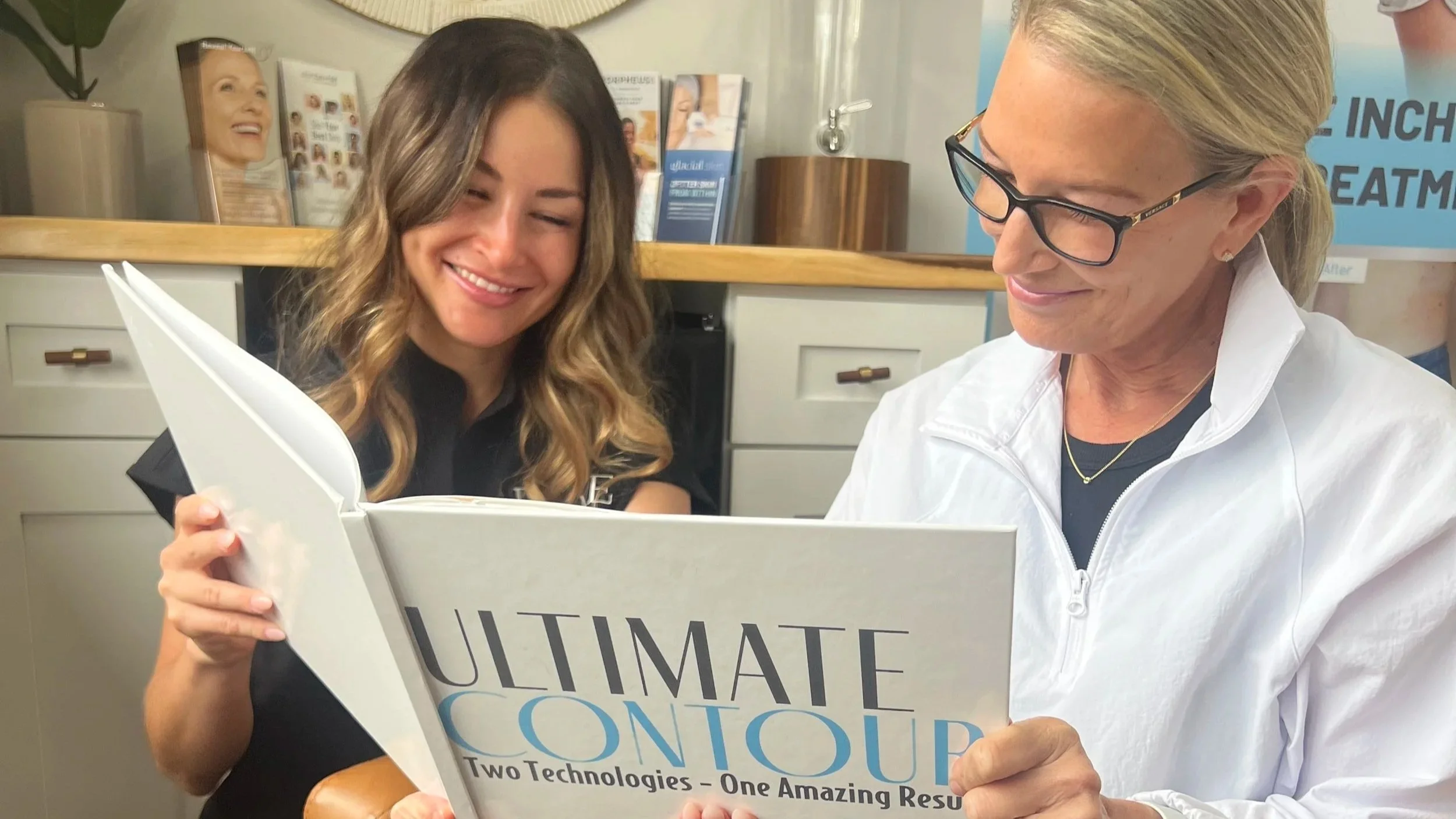 Two women smiling while reading a brochure titled "Ultimate Contour" in a bright office setting. The mood is positive and professional.