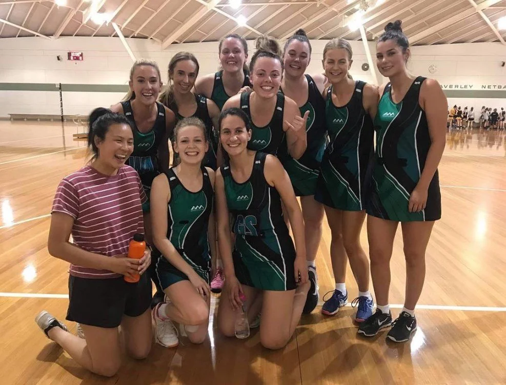 A group of nine young women in tennis uniforms posing together on an indoor basketball court, smiling and celebrating after a game.