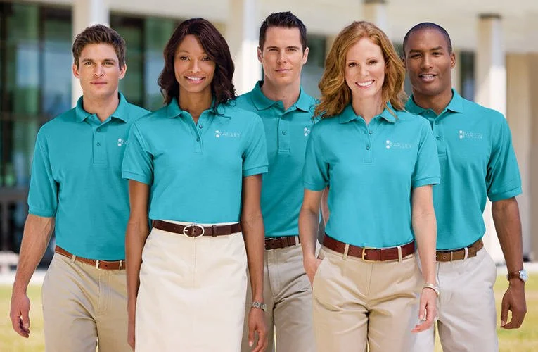 Group of five diverse adults wearing matching teal polo shirts with a company logo, smiling, outside in front of a modern building.