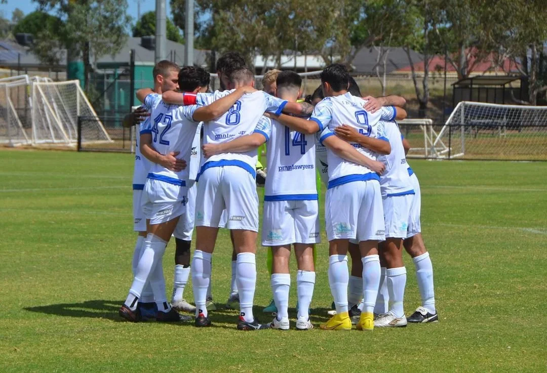 Group of soccer players in white uniforms with blue accents huddled together on a grass field during daytime.