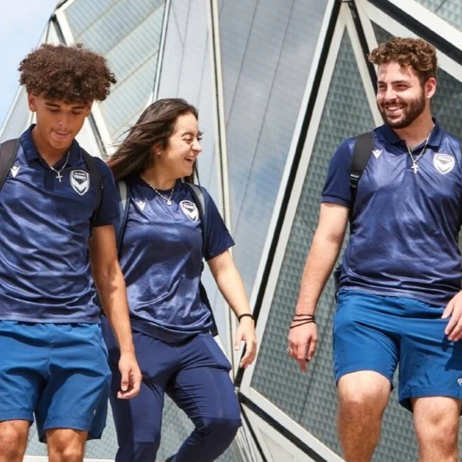 Three young adults wearing matching navy blue sports jerseys and shorts, walking outdoors in front of a modern glass building. They are smiling and seem to be enjoying each other's company.