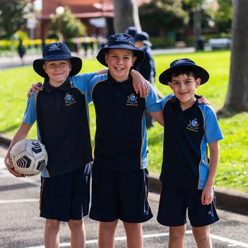 Three young boys in blue and black uniforms with wide-brimmed hats, smiling and standing outdoors on a sunny day, one holding a soccer ball, with trees and a park in the background.