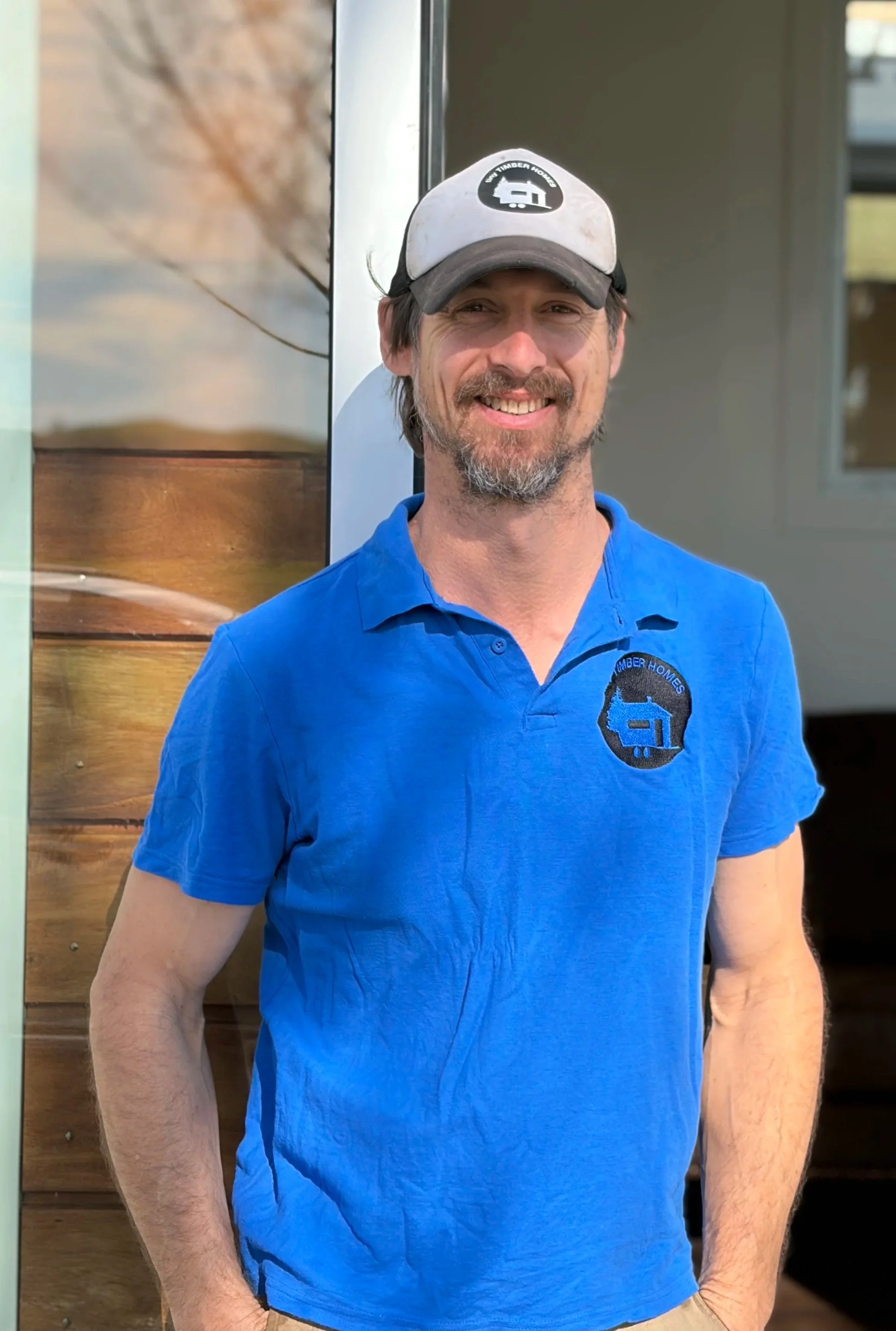 A man sitting on a toilet, wearing a blue polo shirt with a logo, tan shorts with frayed edges, and a white baseball cap. The bathroom has wooden cabinets and a window in the background.