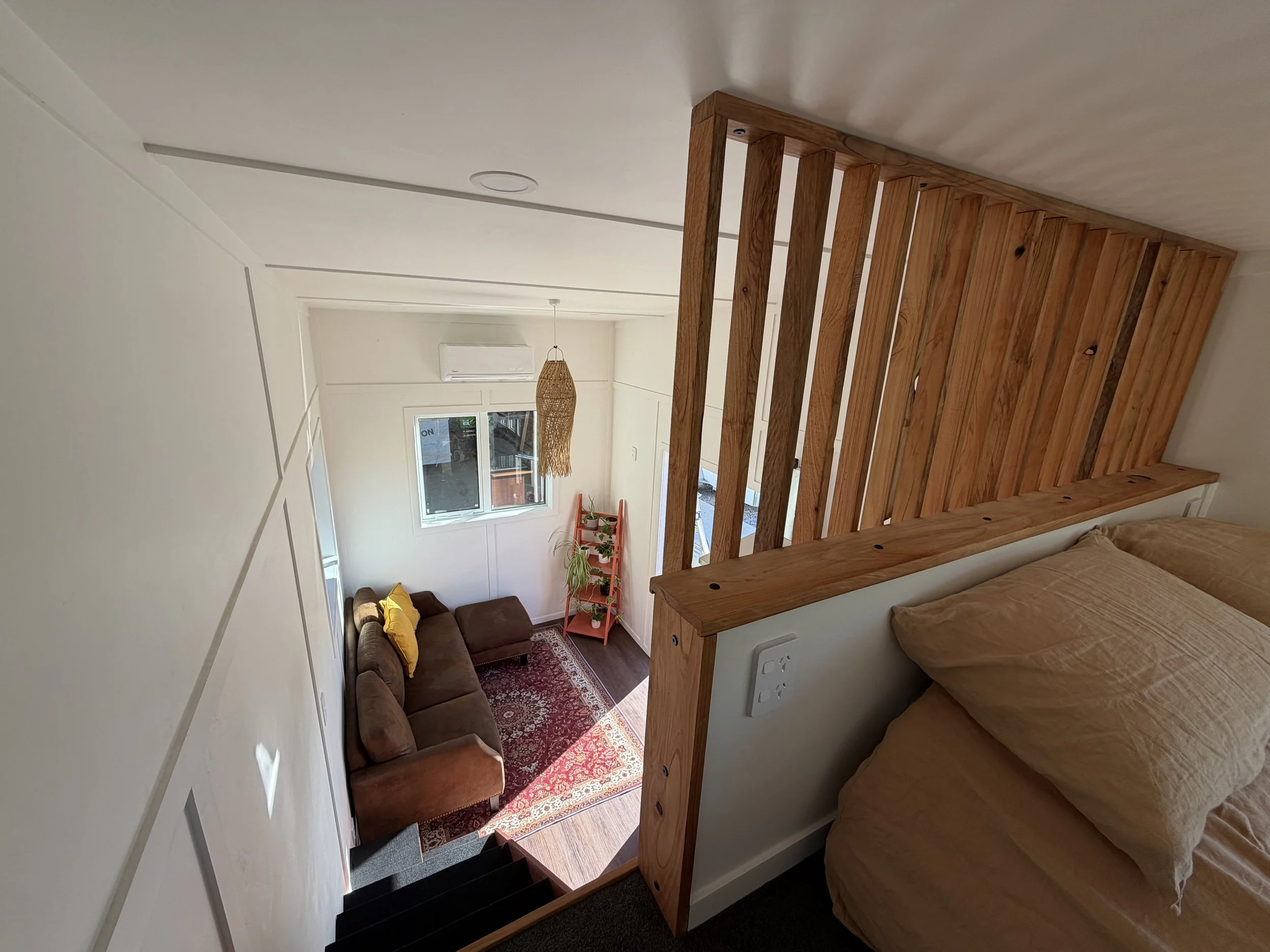 Tiny Timber Homes rental loft bedroom with timber slat railing, overlooking lounge with sofa and heat pump air conditioning