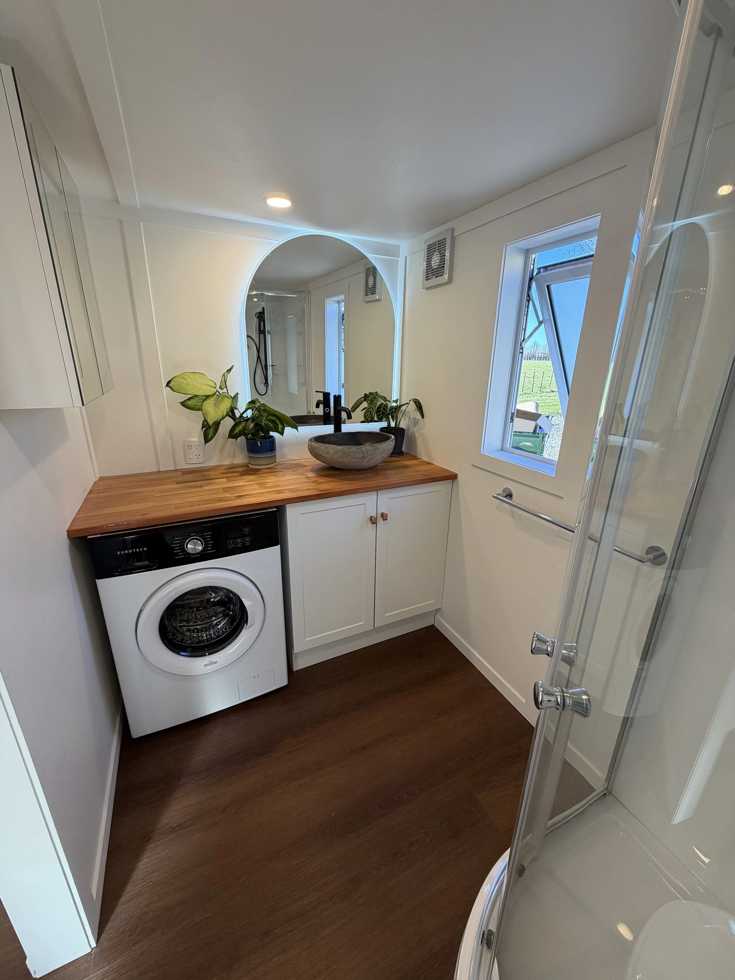 Tiny Timber Homes rental bathroom with washing machine, timber vanity, stone vessel sink, arched mirror, and glass shower door