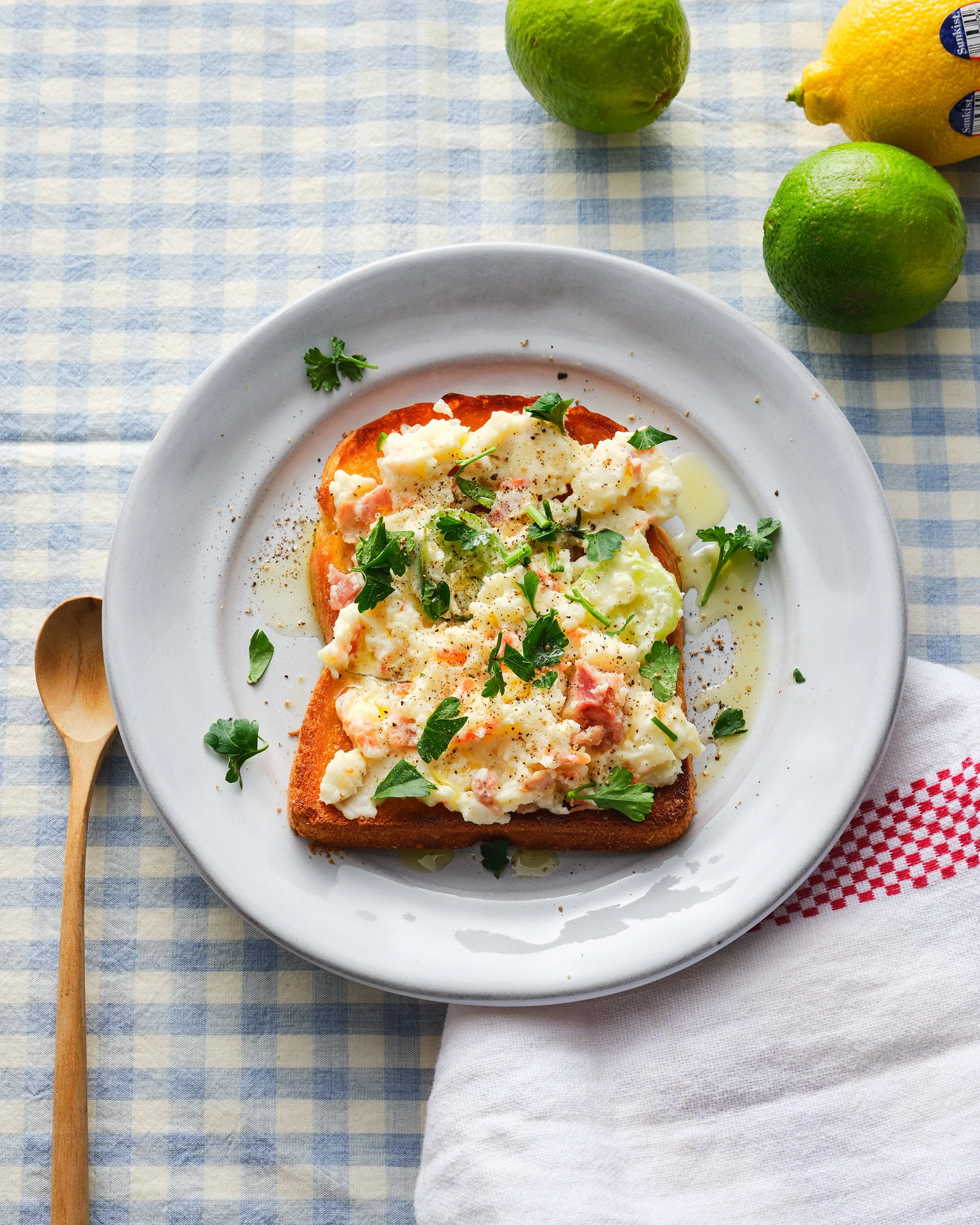 "파란색 체크무늬 식탁보 위에 으깬 감자 샐러드와 연어를 듬뿍 올린 토스트가 하얀 접시에 담겨 있습니다. 라임, 레몬, 나무 숟가락이 함께 놓여 소풍 같은 분위기를 연출한 밝고 따뜻한 브런치 푸드 스타일링 사진입니다."