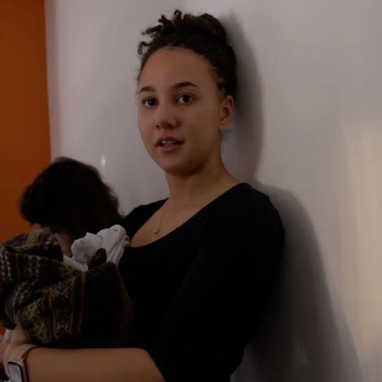 Young woman with curly hair holding a small dog and standing against a white wall.