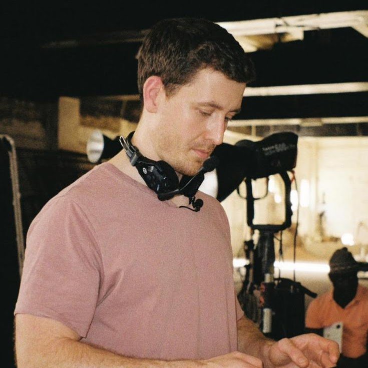 A man with headphones around his neck looking down at his hands, on a film set or photo shoot, with equipment and another person in the background.