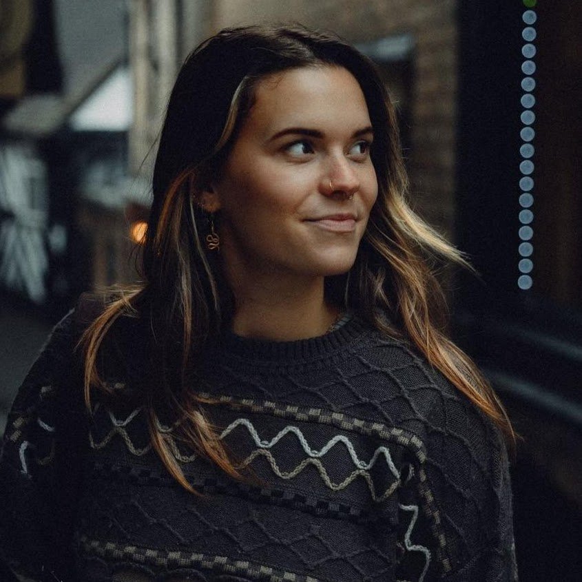 A young woman with long, wavy hair and a nose ring, wearing a patterned sweater, looking to her left in a dimly lit indoor setting.