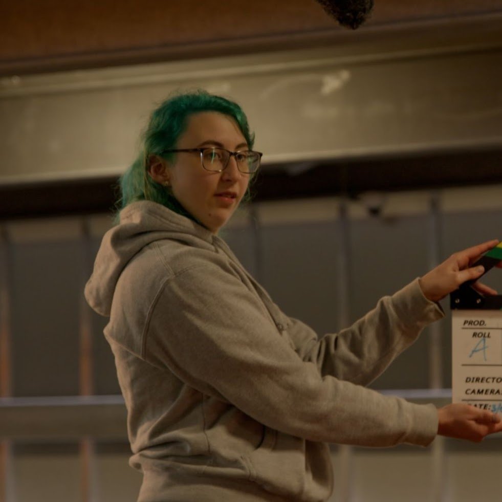 Young woman with green hair and glasses holding a clapperboard.
