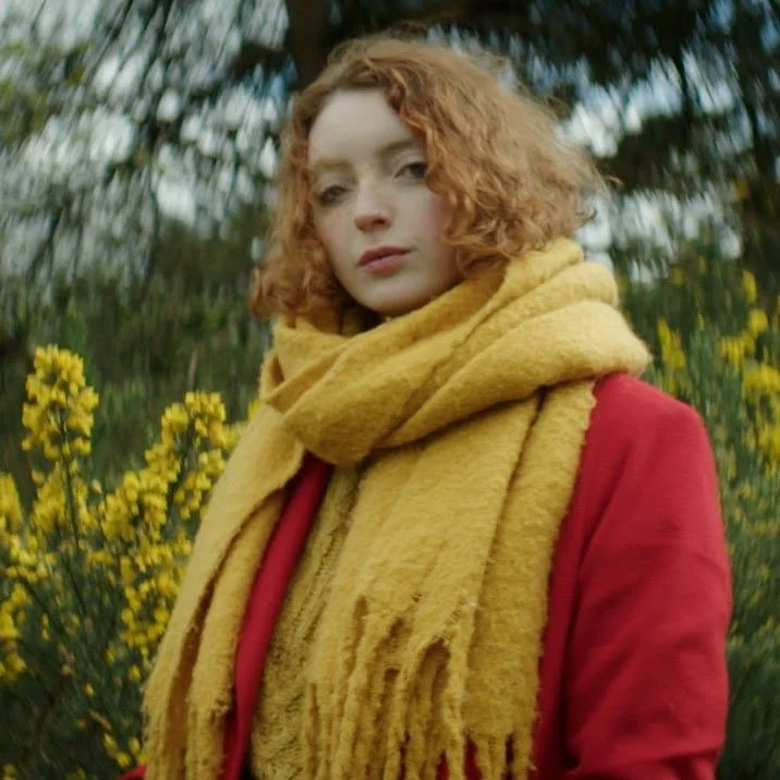 A woman with curly red hair wearing a yellow scarf and red jacket standing outdoors near yellow flowers and green trees.