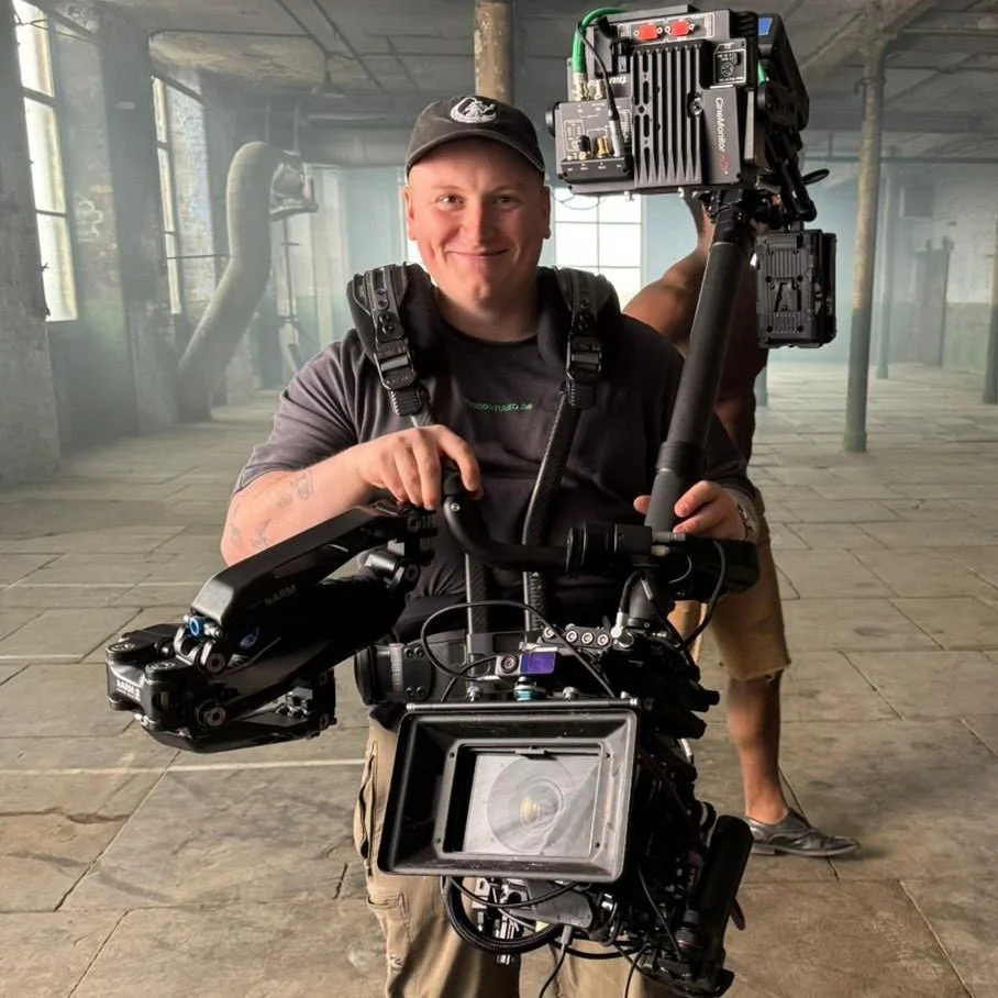 A smiling man wearing a cap and black t-shirt holding a professional camera rig on a Steadicam stabilizer inside an industrial space with large windows and exposed pipes.