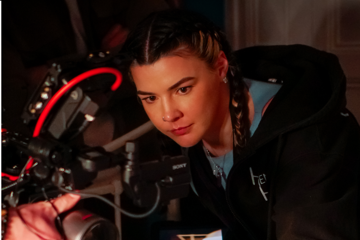 A young woman with braided hair and a nose piercing watching a film crew camera.