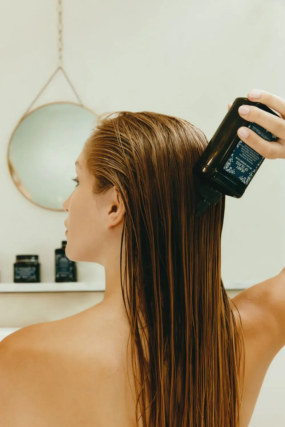 A woman with long, wet, red hair is using a spray bottle to apply hair treatment in a bathroom with a mirror and shelves with black bottles.