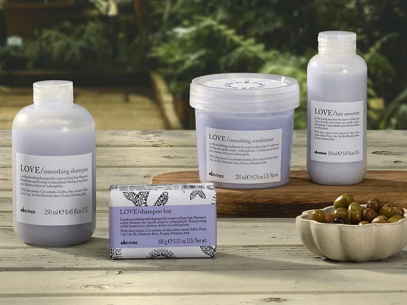 Assorted hair care products on a wooden surface with a bowl of green and brown olives nearby and a blurred outdoor background.