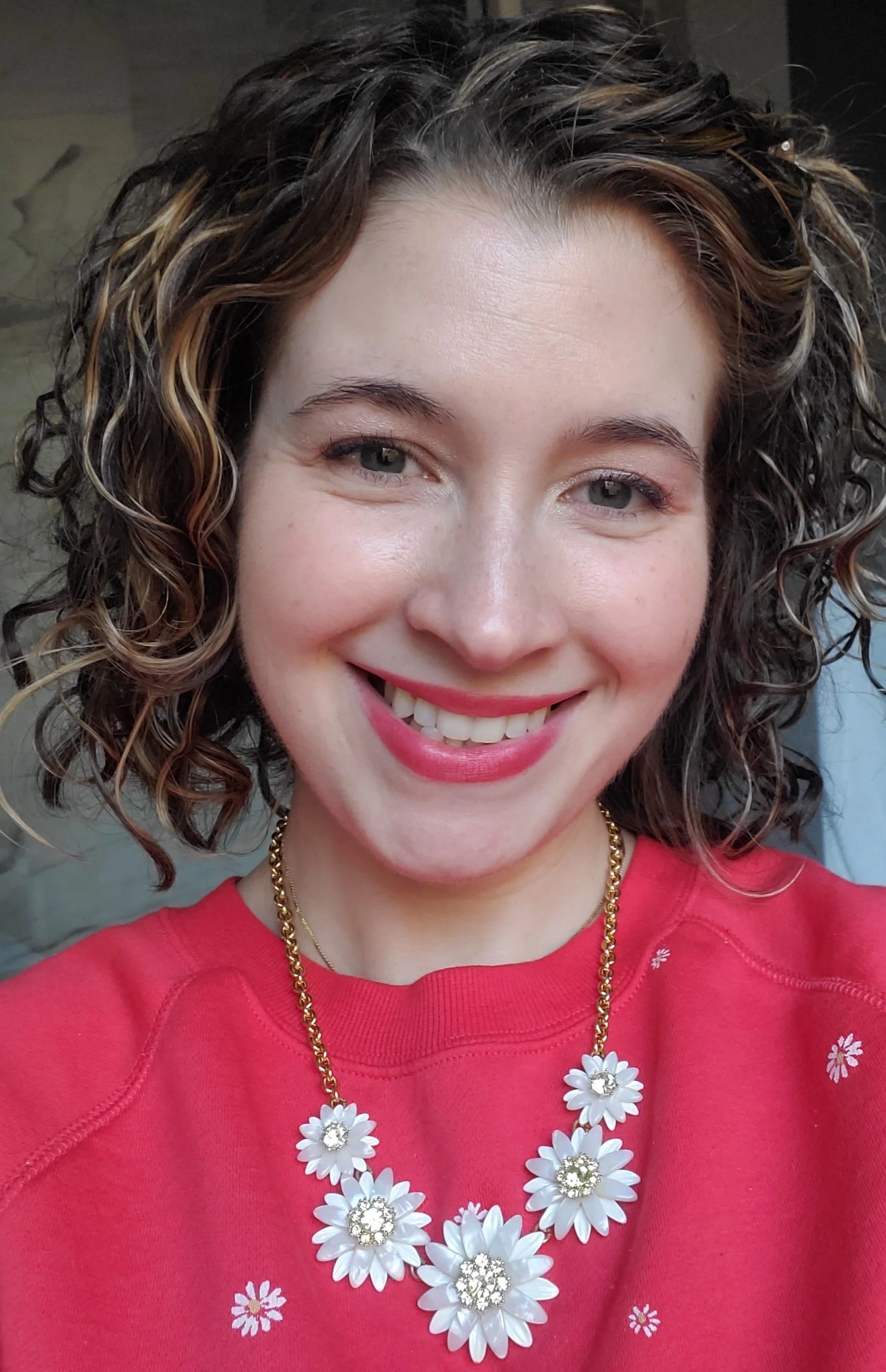 Person smiling, wearing a red top and a daisy-themed necklace.