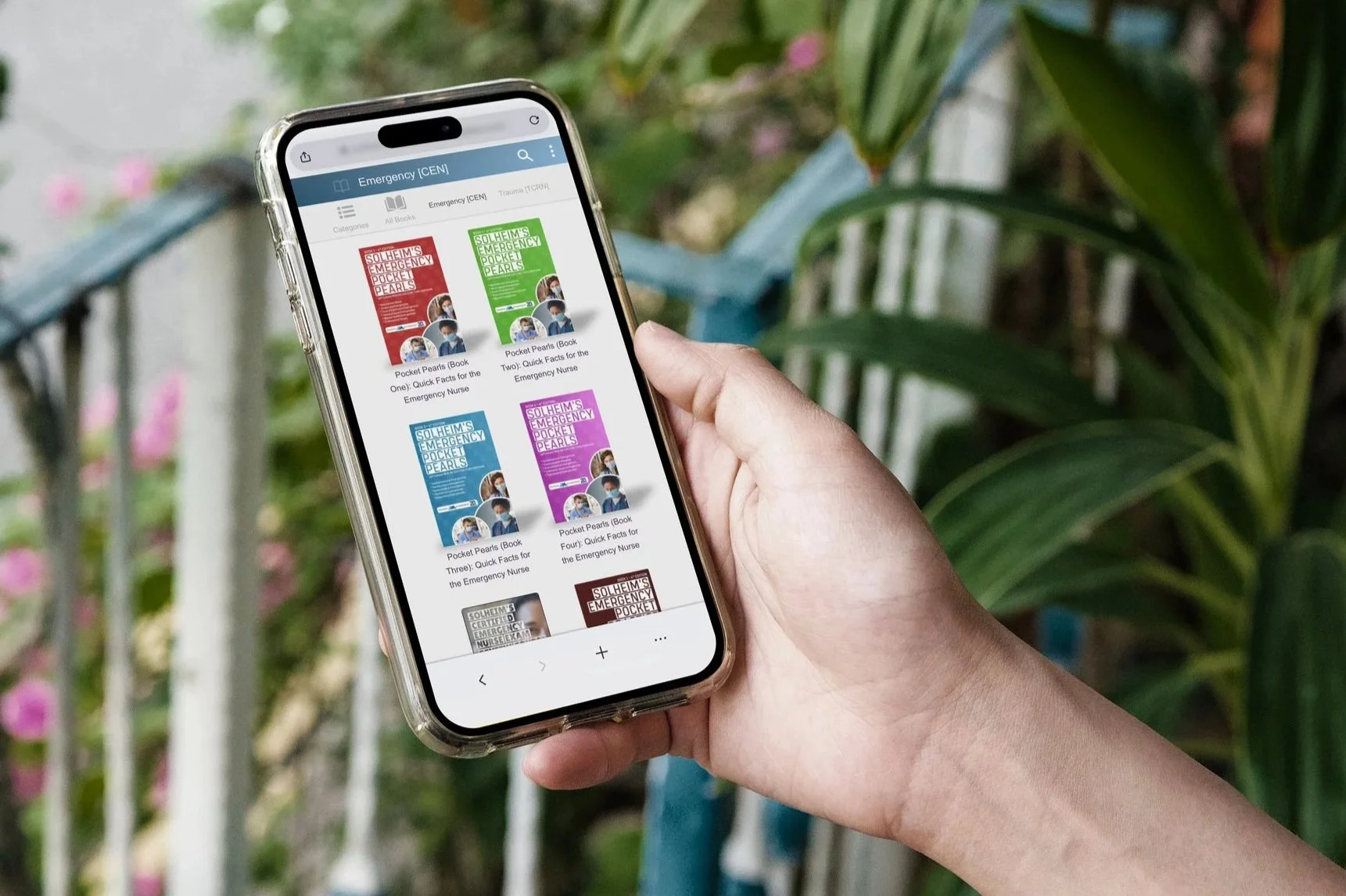 A person holding a smartphone displaying various emergency pocket guidebooks in digital format, with a blurred background of green plants and a picket fence.