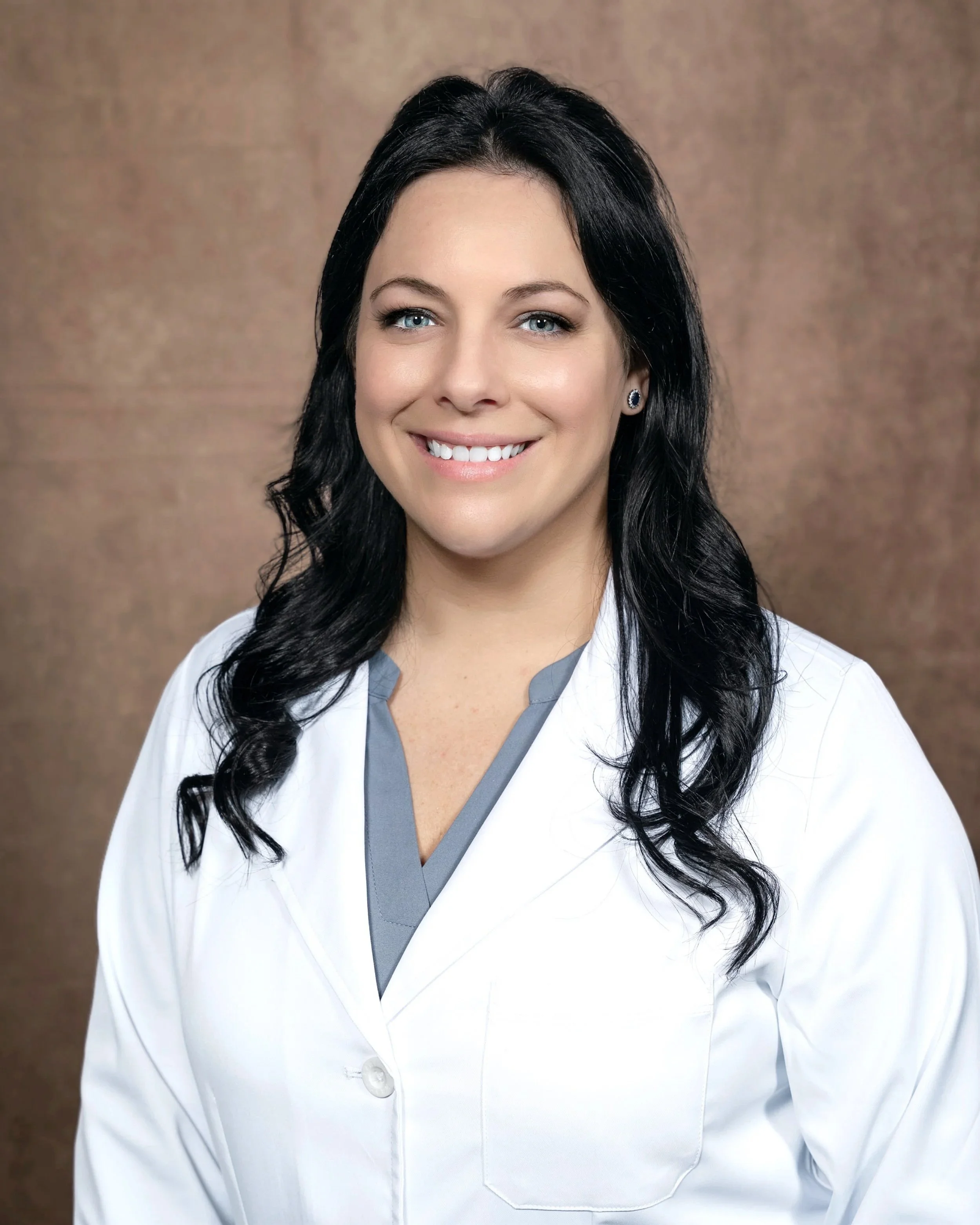 A woman with black hair, blue eyes, and light skin wearing a white medical coat with grey scrubs underneath, smiling and looking at the camera, standing in front of a brown textured background.