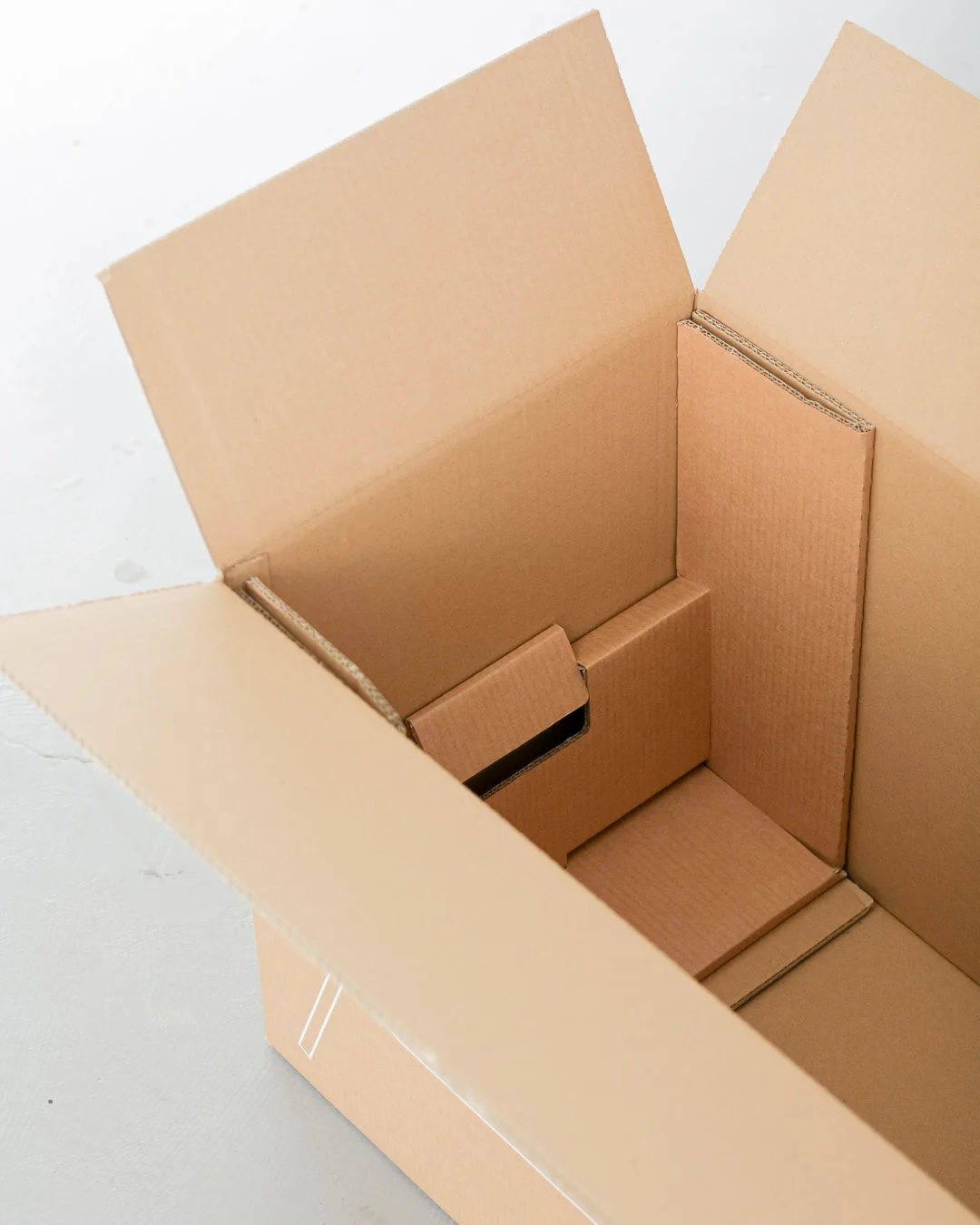 Empty brown corrugated cardboard box, open at the top, with its flaps folded outward, placed on a white surface.