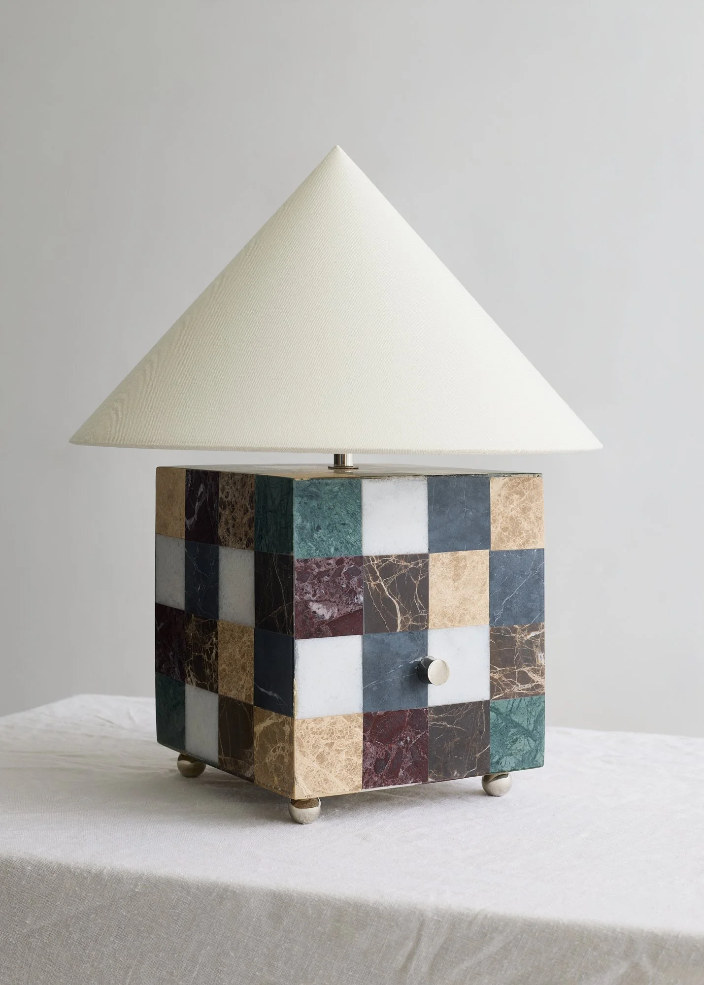 Angled view of The Crews table lamp showing two sides of its checkerboard marble base beneath an ivory fabric shade.
