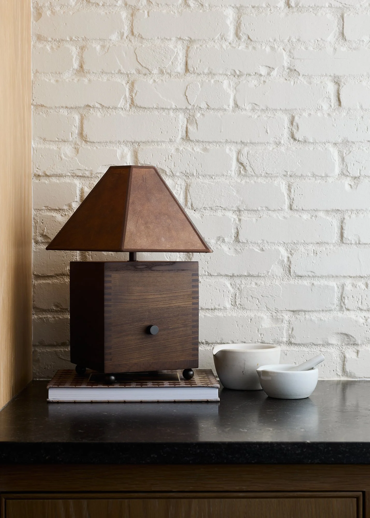 The Carley table lamp styled on stacked books beside white ceramic bowls against a white brick wall.
