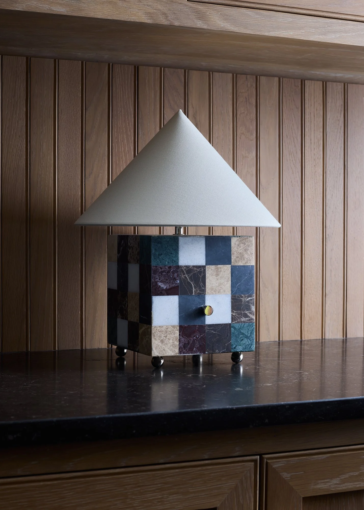 The Crews specimen marble table lamp on a dark tabletop against wood paneling, with a pale fabric shade and polished nickel feet.