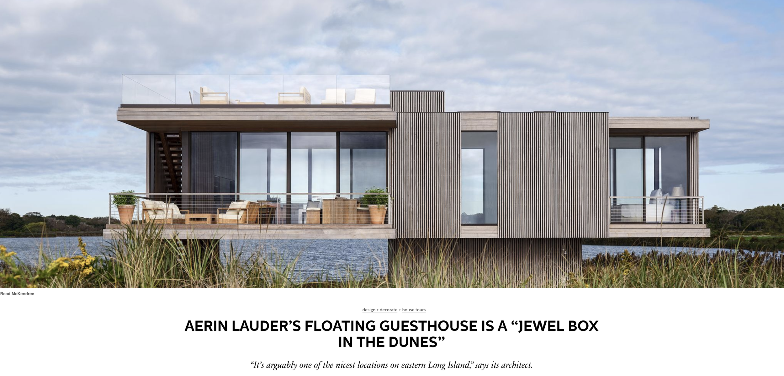 Modern floating house with large glass windows, outdoor balcony, and wooden exterior, situated on a body of water surrounded by grass and shrubbery in Sagaponack New York