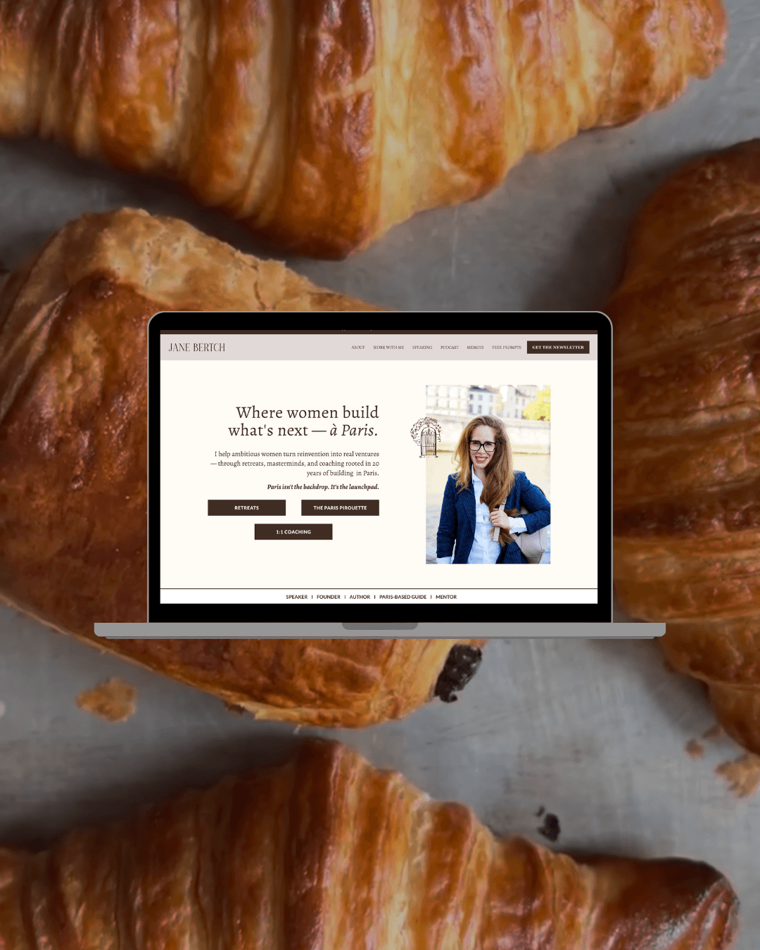 Laptop displaying a website with the text 'Where women build what's next — à Paris', surrounded by close-up images of croissants.