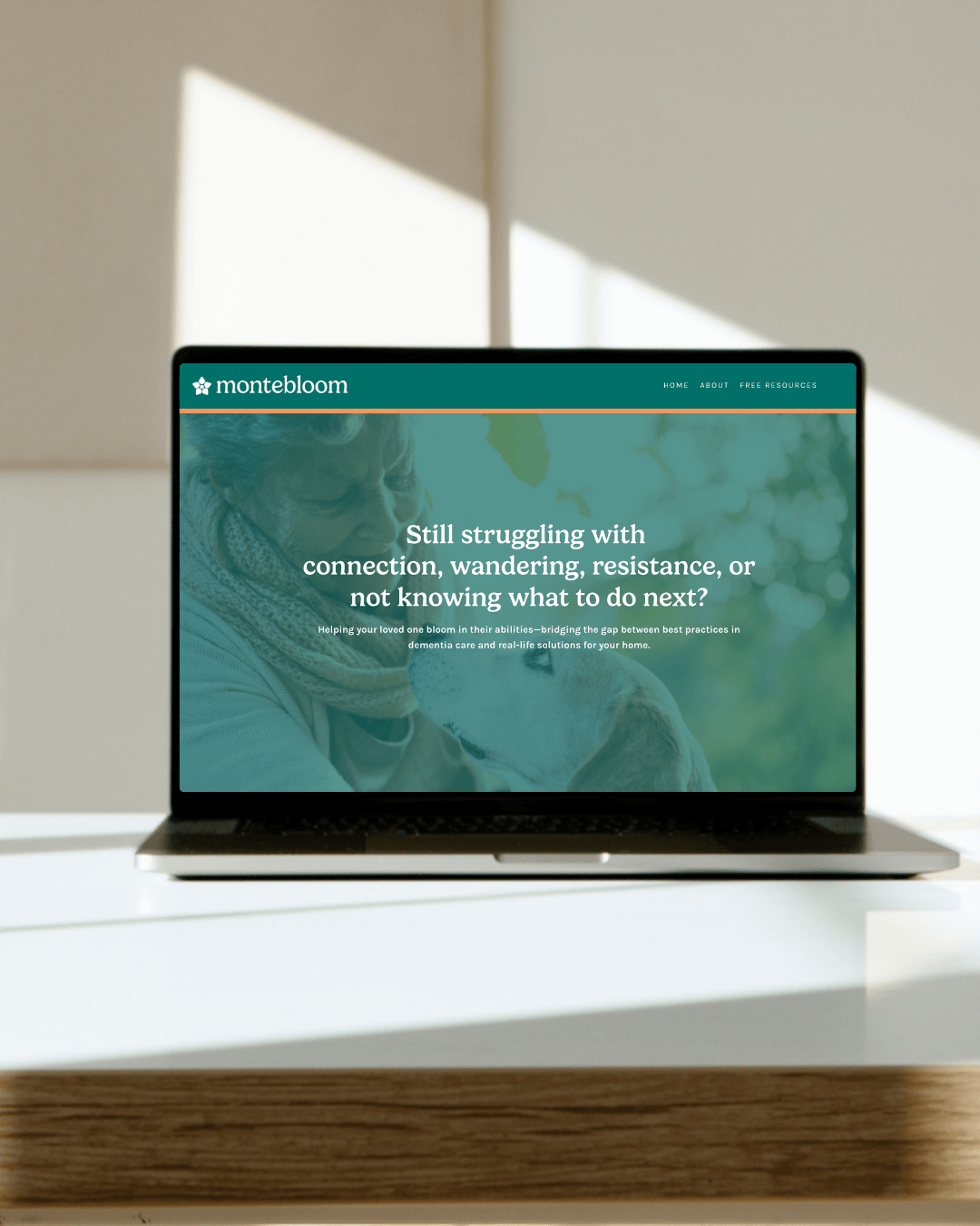 Laptop screen displaying the Montebloom website with a background image of an elderly woman with a dog, sitting outdoors with green foliage, in a bright room with sunlight and a window casting shadows.