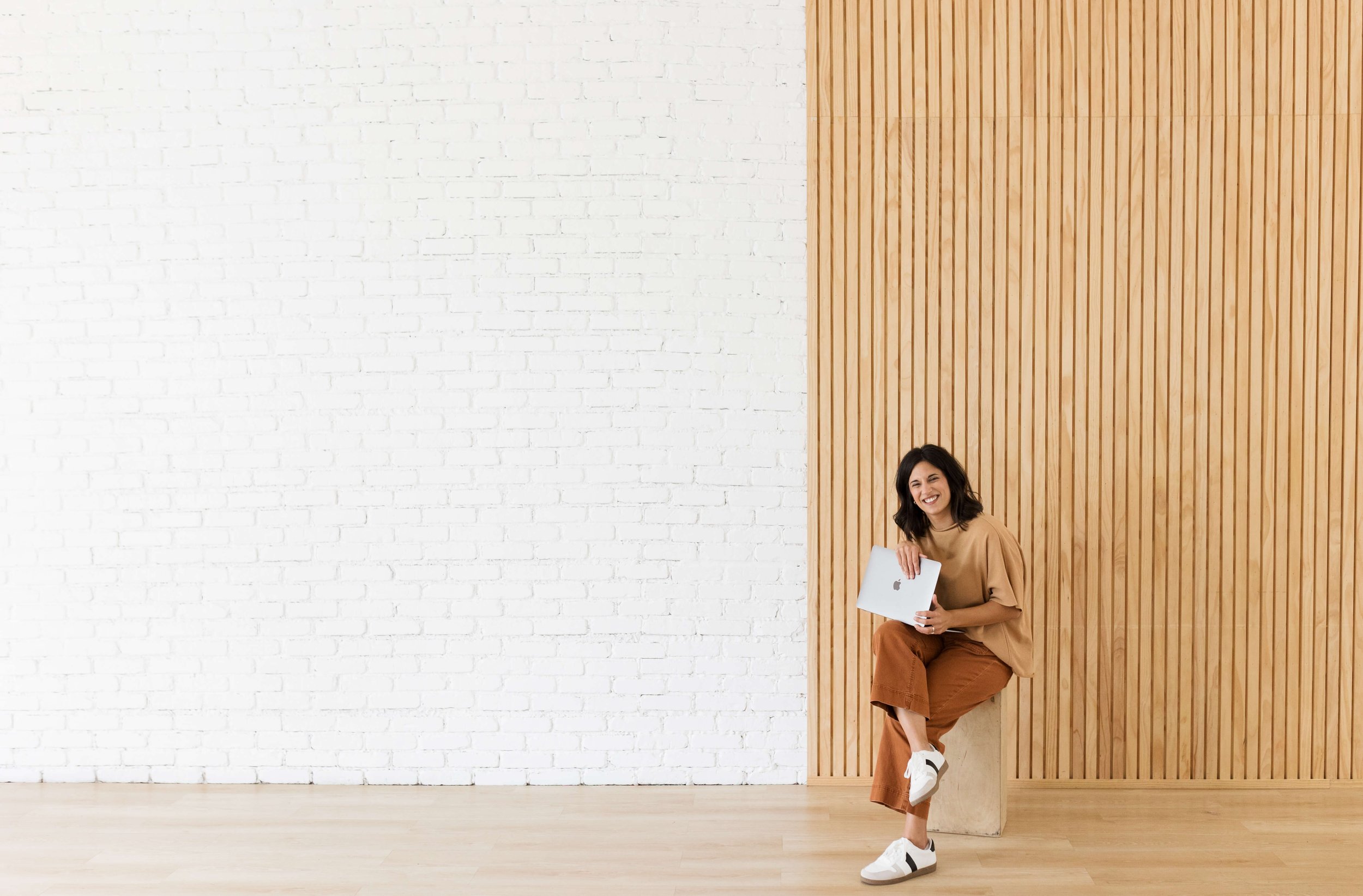 Woman smiling while sitting on a stool, holding a laptop, with a white brick and wooden panel wall in the background.