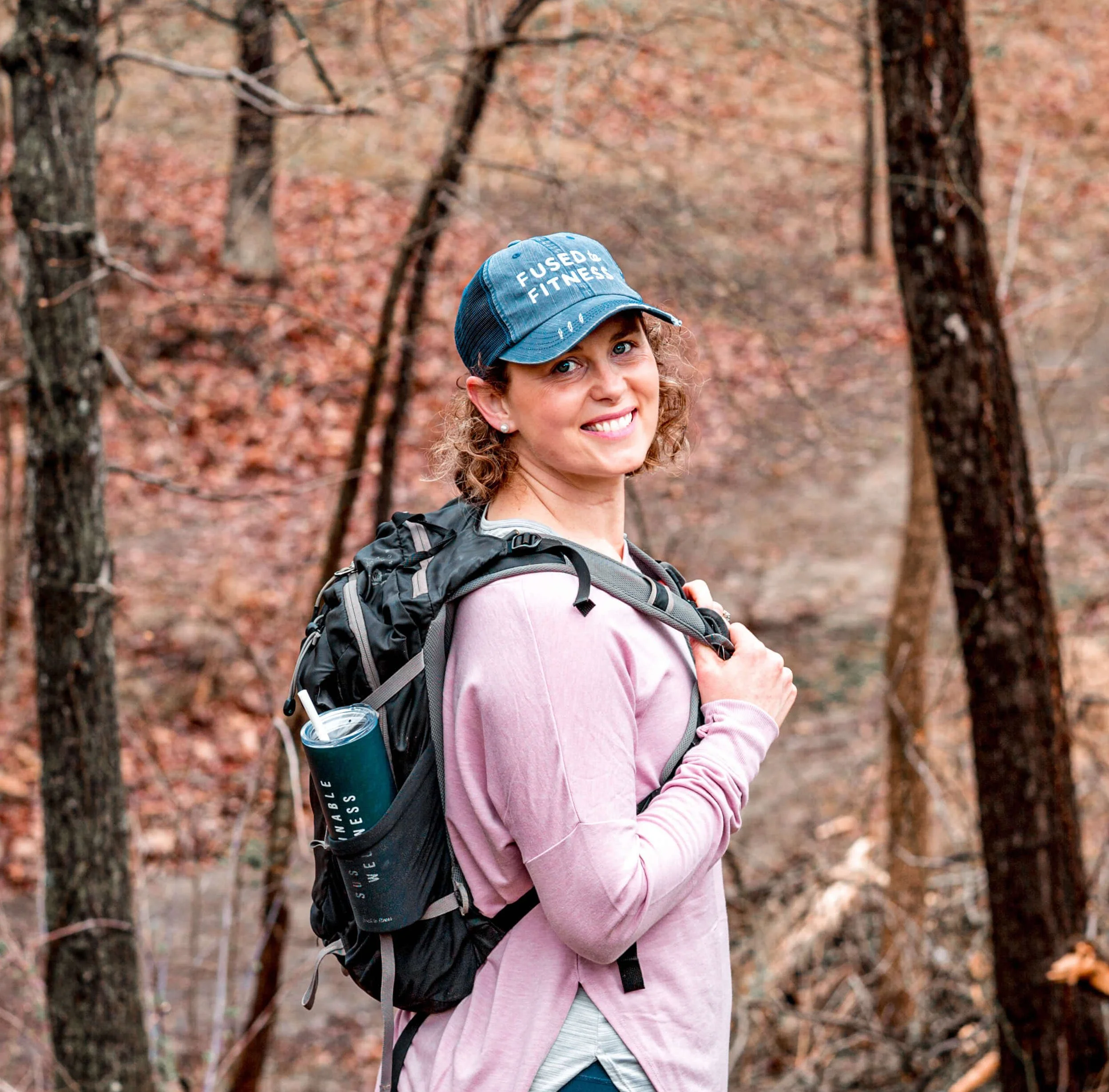 A person in a pink shirt and blue cap hiking on a wooded trail, carrying a backpack with a water bottle.