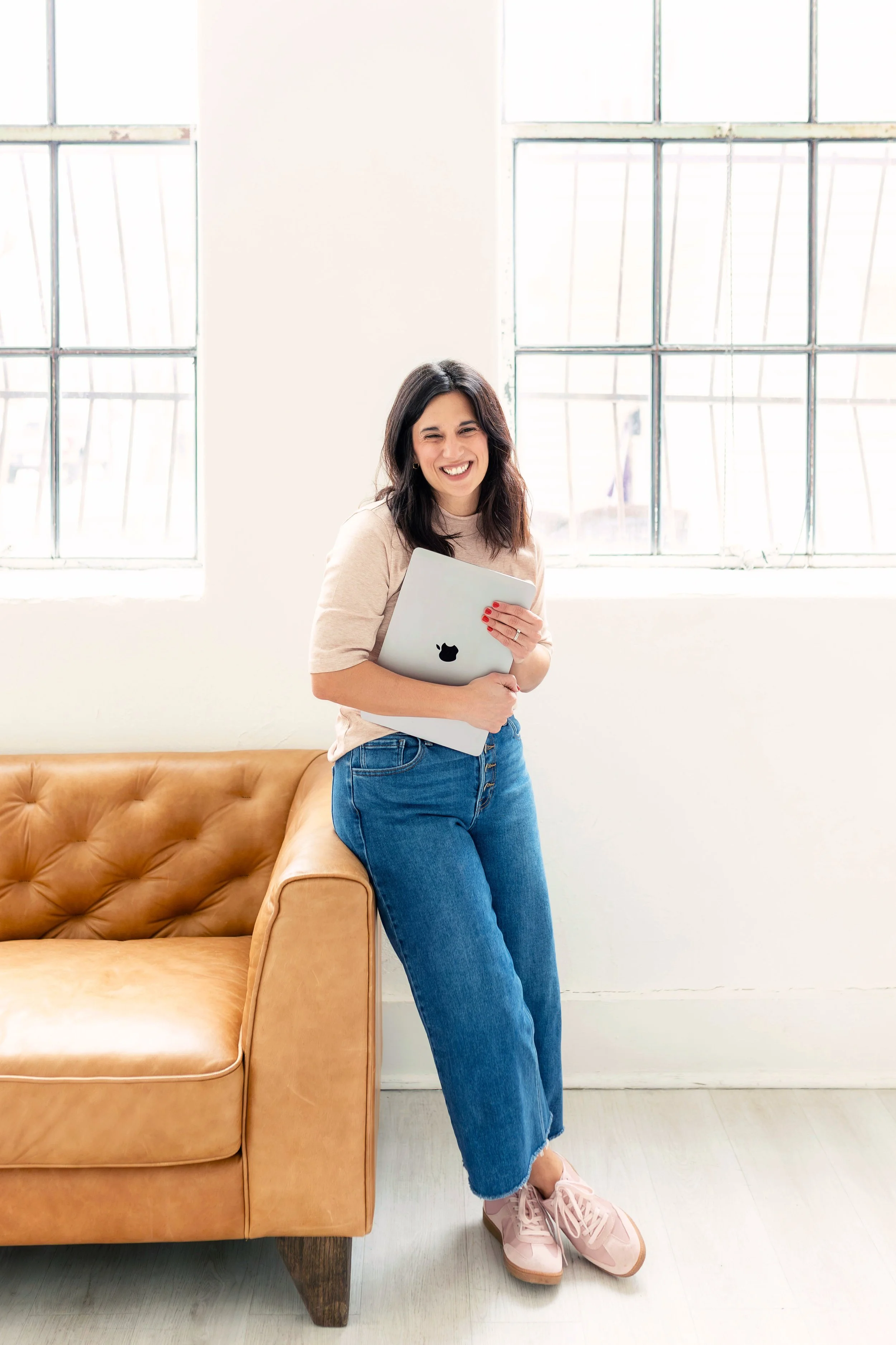 A woman standing next to a tan leather sofa, smiling and holding a closed MacBook, inside a bright room with large windows.