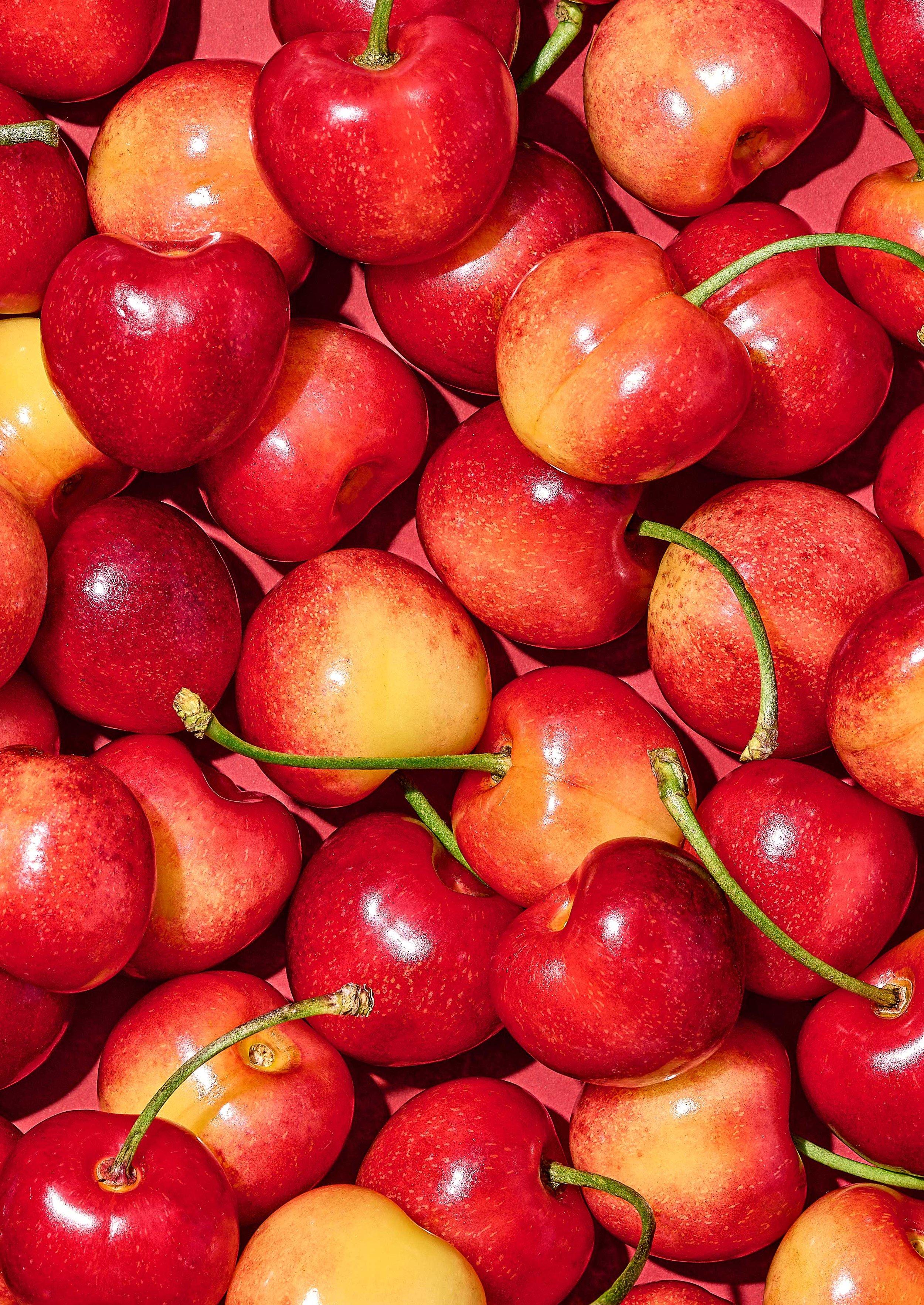 Overhead macro shot of Rainier cherries with their signature pink-and-gold blush, densely arranged on a coral red backdrop for Suzanne Clements' Publix summer produce campaign.