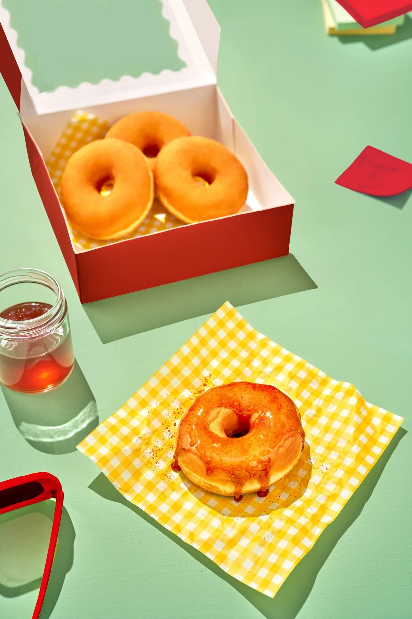 Commercial food photography by Suzanne Clements, The Sauce Boss spec campaign: a plain donut glazed in chili-flecked hot honey on yellow gingham paper, beside a red bakery box of donuts and a honey jar on a mint table in hard sunlight.