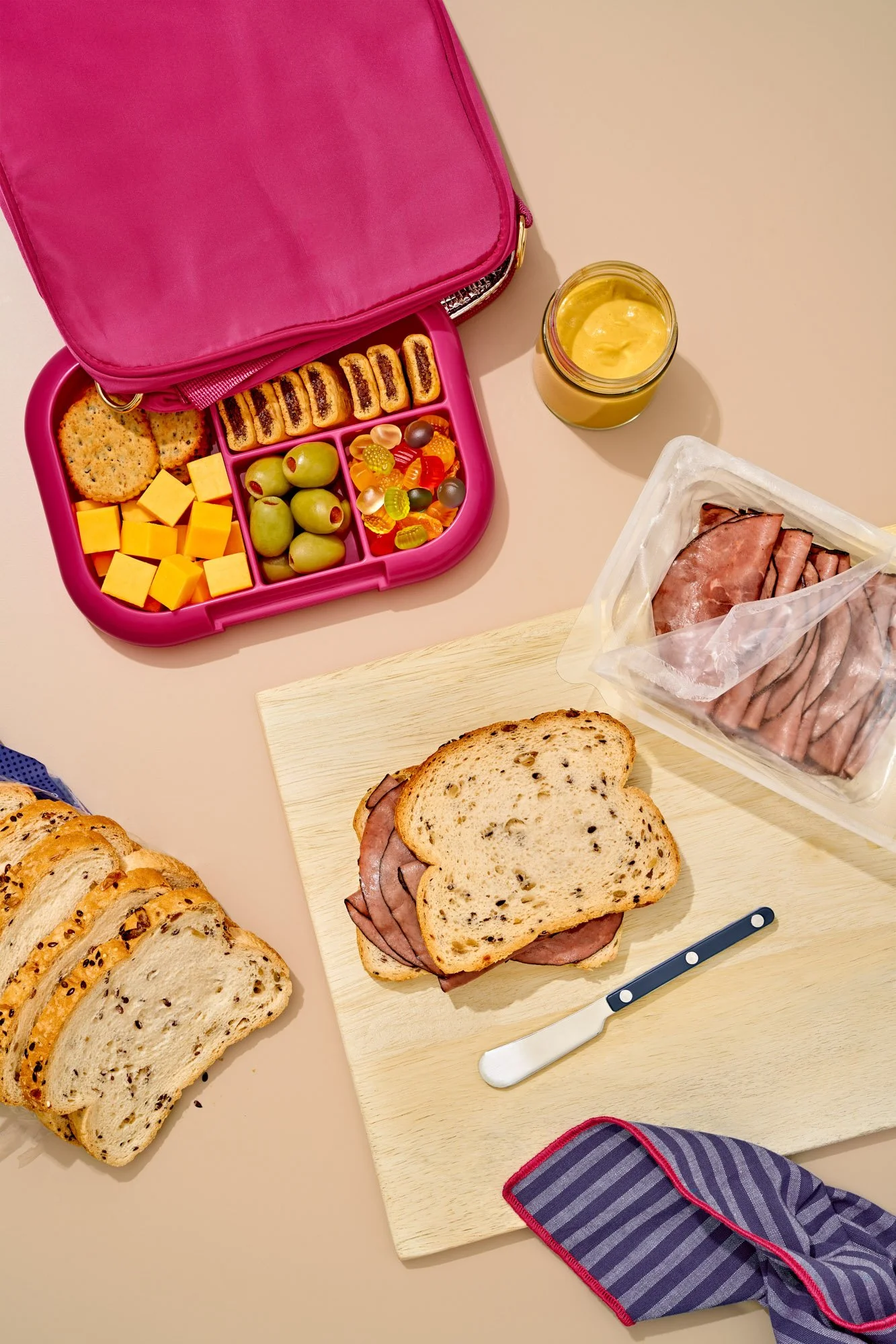 Commercial food photography by Suzanne Clements created for Non UPF certification. This image features a lifestyle food prep moment with a full bento lunchbox filled with tasty snacks, a loaf of seeded bread, and some lunchmeat ready to wrap up prep 