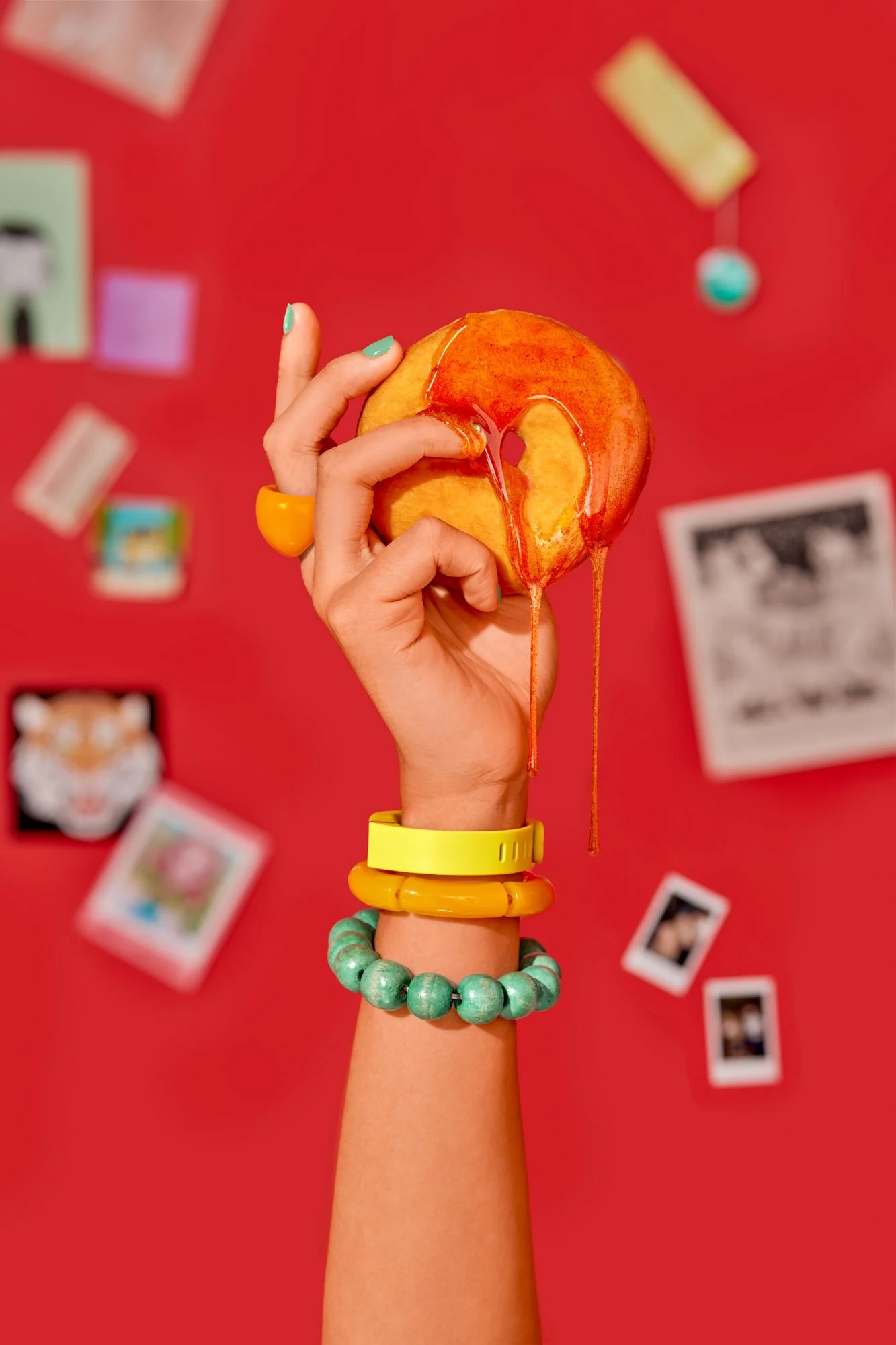 Commercial food lifestyle photography by Suzanne Clements, The Sauce Boss spec campaign: a raised hand grips a donut as glossy chili hot honey runs over the fingers, with stacked yellow and teal bracelets against a red collage wall.
