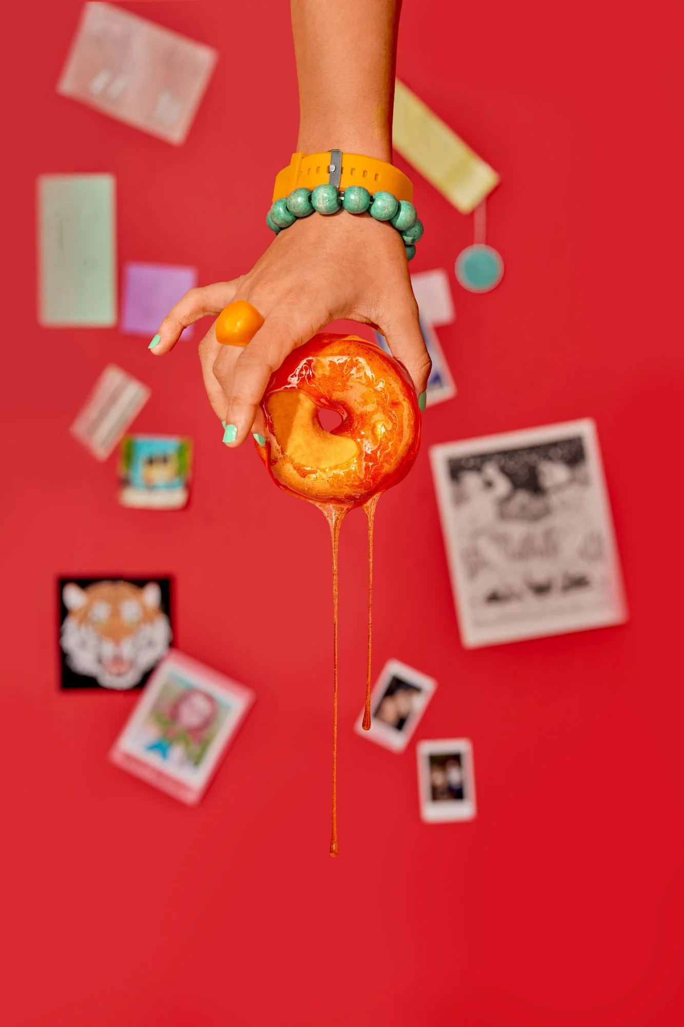 Commercial food lifestyle photography by Suzanne Clements, The Sauce Boss spec campaign: a hand with mint nails, a chunky orange ring, and a teal bead bracelet dangles a donut trailing two long hot honey drips over a red wall of dorm ephemera.