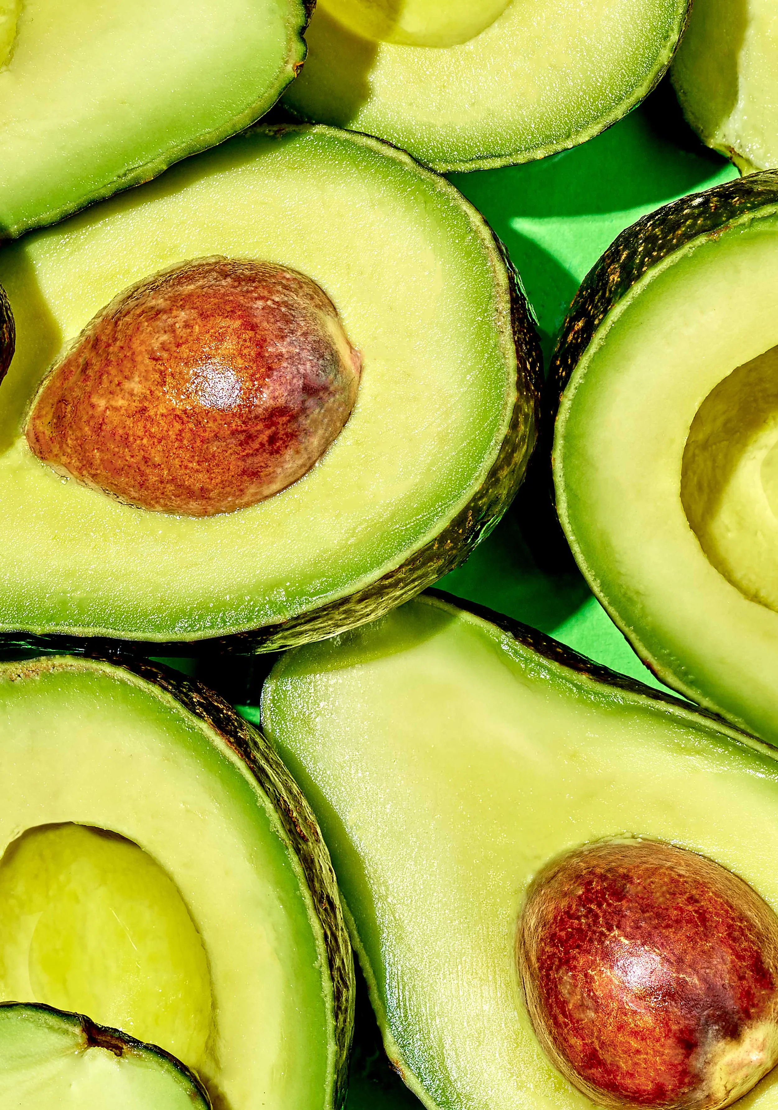 Overhead macro shot of halved avocados with visible pits, arranged on a bright green backdrop for Suzanne Clements' Publix summer produce campaign.