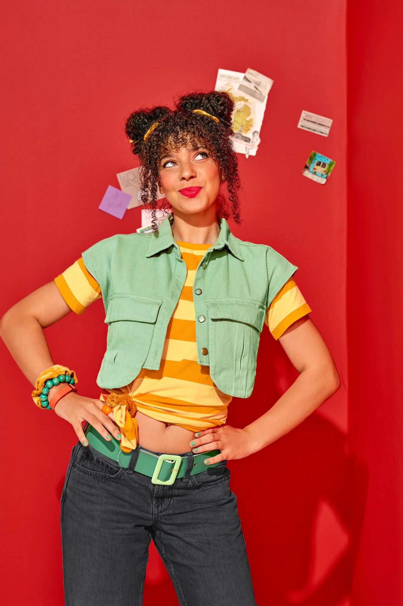Commercial lifestyle photography by Suzanne Clements, The Sauce Boss spec campaign: a college-aged woman in a mint vest and orange striped tee stands hands on hips, glancing up with a sly smirk in front of a red wall pinned with ephemera.