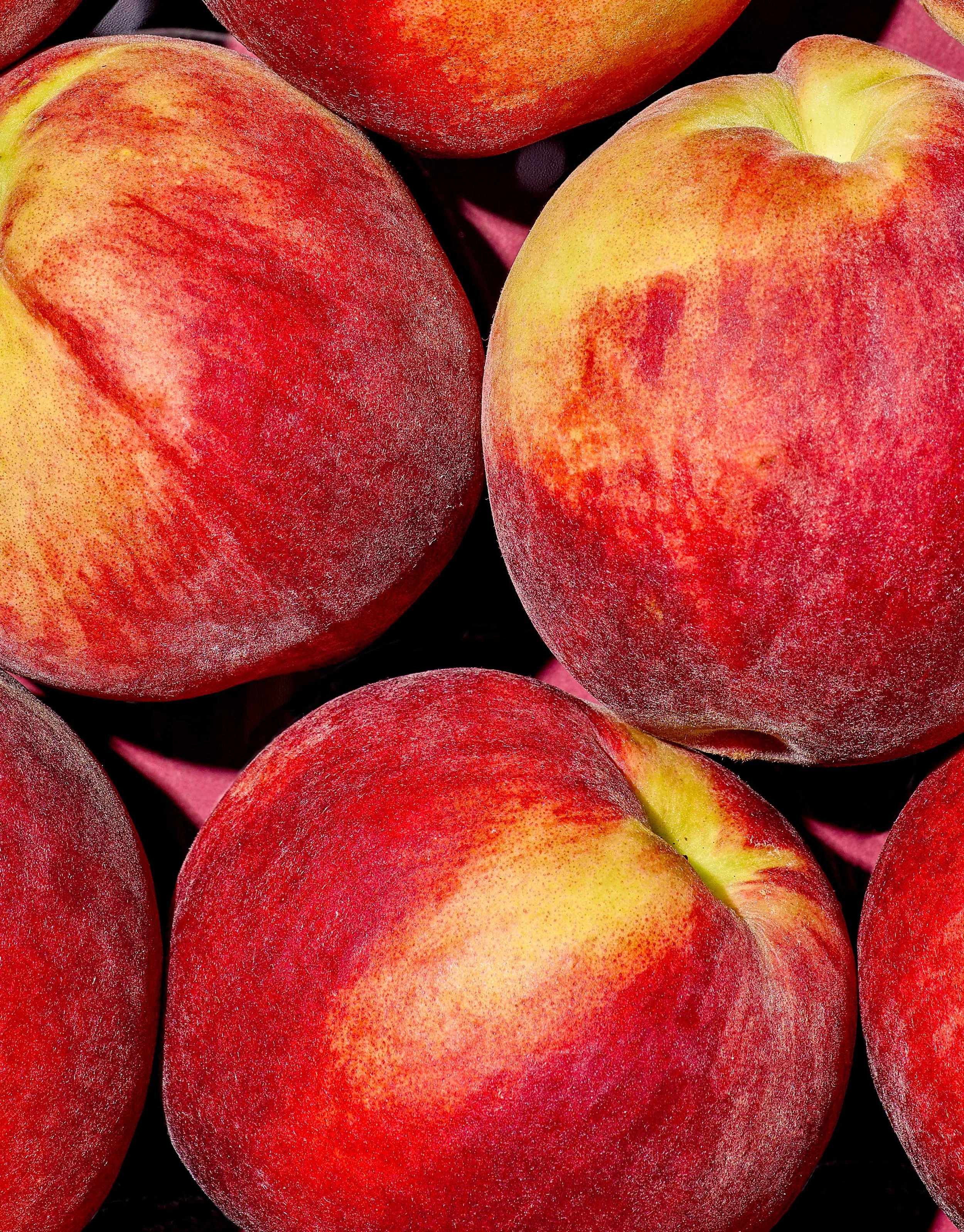 Overhead macro shot of whole peaches with red and gold fuzzy skin, densely arranged on a deep rose backdrop for Suzanne Clements' Publix summer produce campaign.