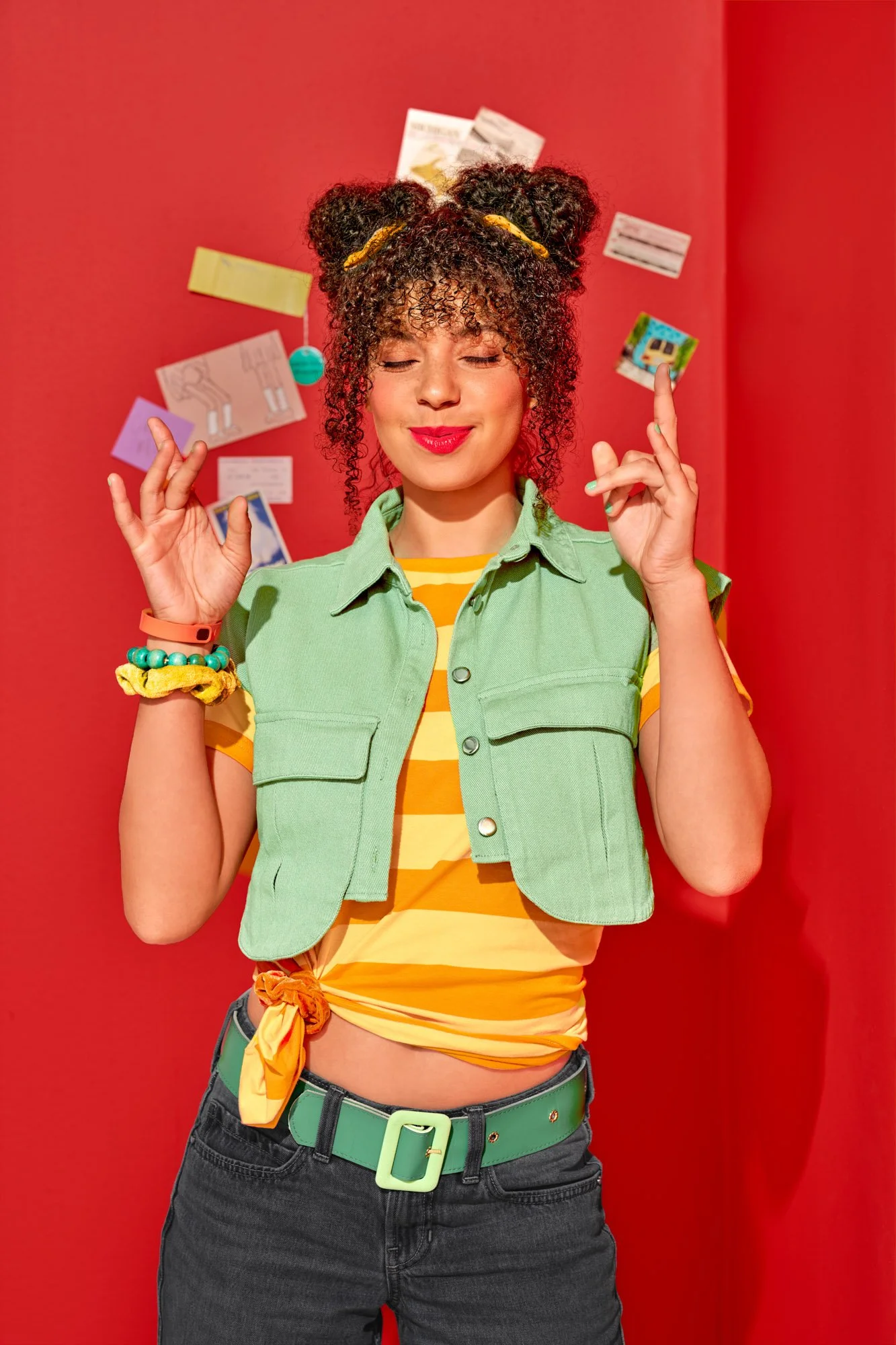 Commercial lifestyle photography by Suzanne Clements, The Sauce Boss spec campaign: a college-aged woman with curly space buns, a mint cropped vest, and a striped tee closes her eyes and crosses her fingers against a red wall.